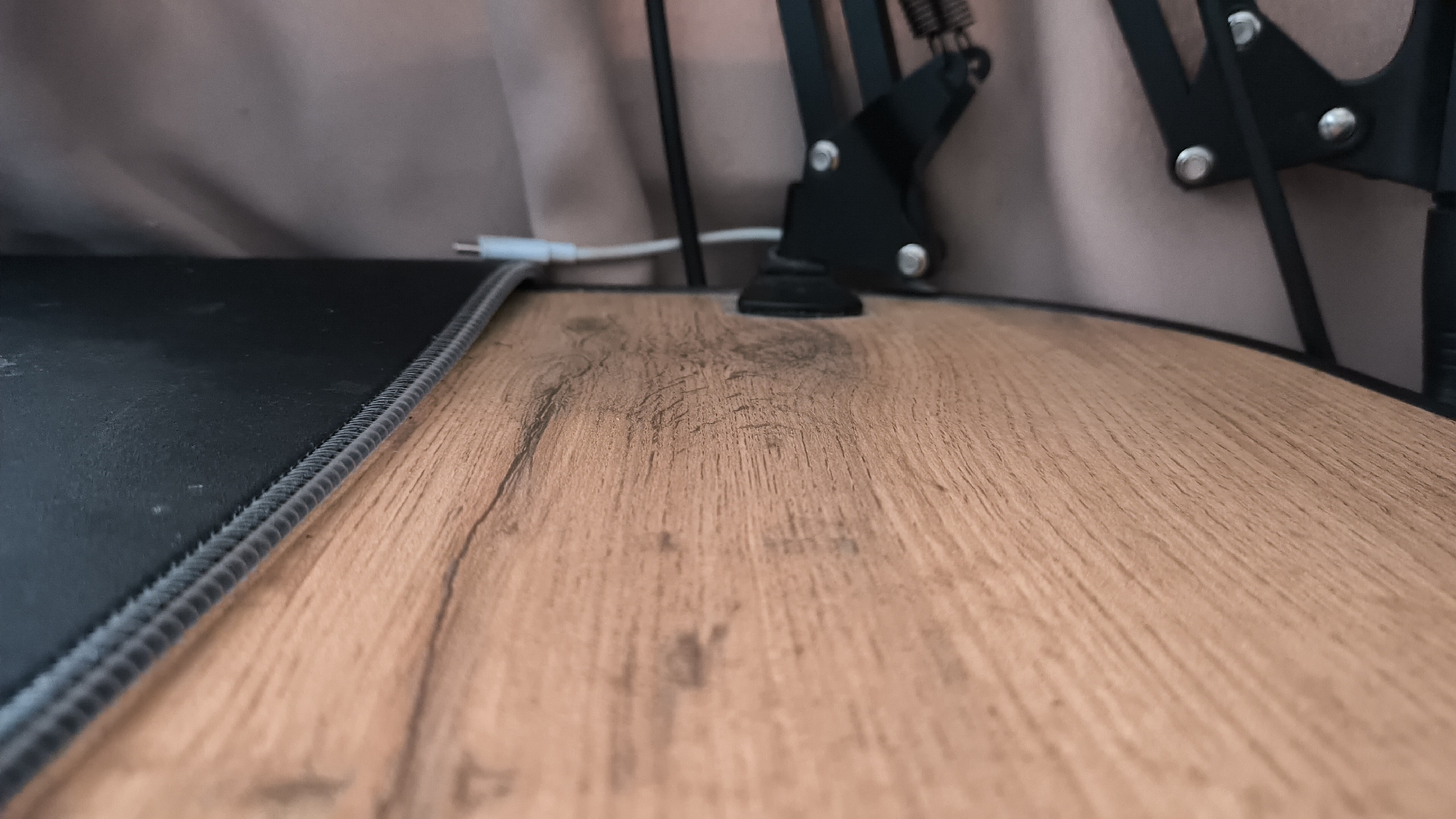 The image shows a low-angle, close-up view of a wooden surface with a black mousepad on the left. The wooden surface has a visible grain pattern. In the background, a black microphone stand is partially visible, along with a white cable connected to it. The environment appears to be indoors, possibly a desk setup. There is no discernible location within the city of Kovrov, Russia, or any specific details about people or actions.
