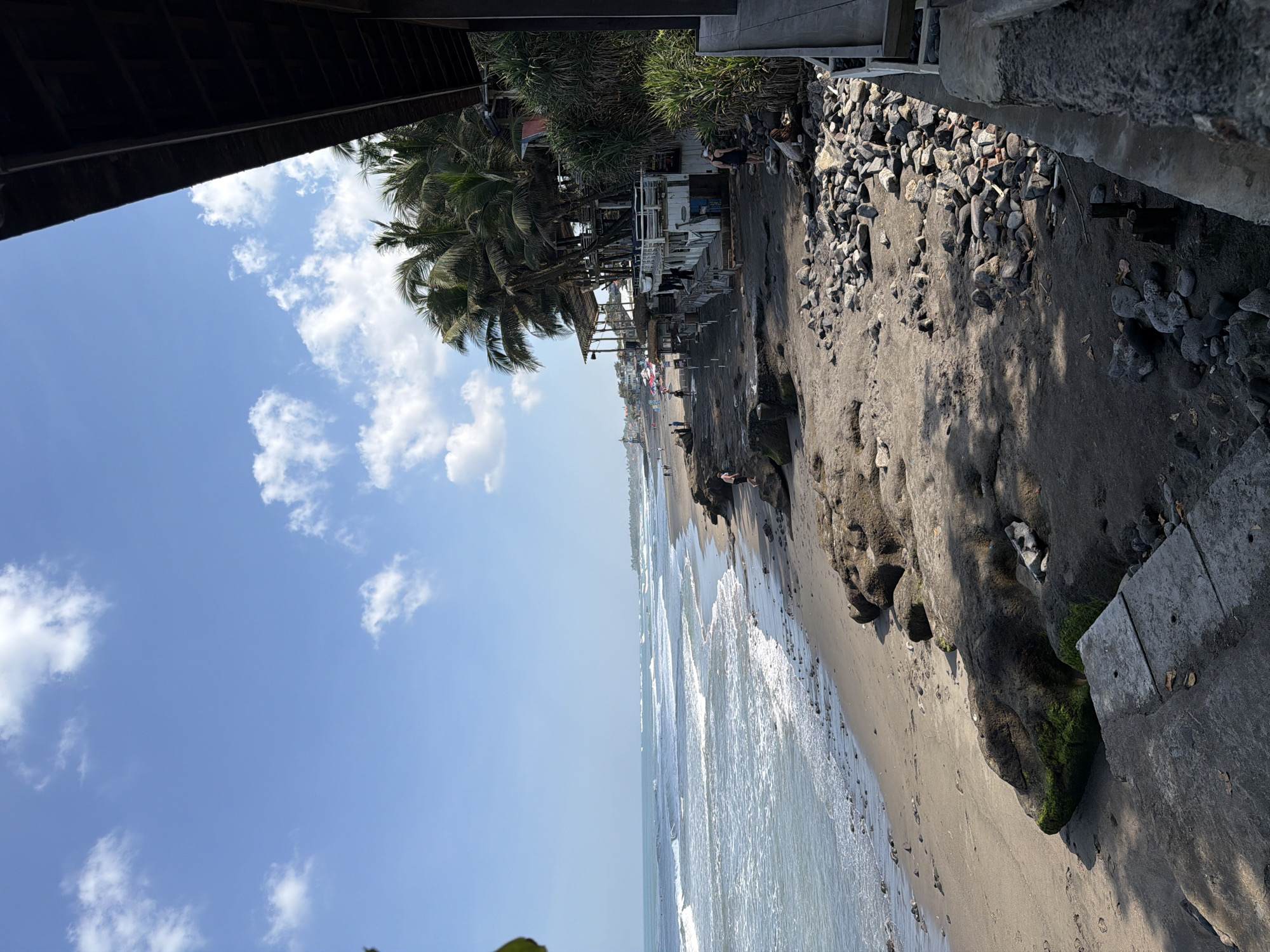 The image depicts a tropical beach scene in North Kuta, Indonesia, under a bright blue sky with scattered white clouds. The foreground features a rocky, dark sand beach with scattered stones and large, moss-covered rocks. A concrete structure or walkway is visible in the lower left corner. Beyond the rocks, the sandy beach extends, with a few people walking and sitting. The ocean is calm with gentle waves rolling towards the shore. Further down the beach, there are several open-air structures, possibly cafes or shops, with thatched roofs and wooden platforms, shaded by numerous palm trees. Some individuals are visible around these structures. The overall atmosphere is relaxed and scenic, indicative of a coastal leisure area.