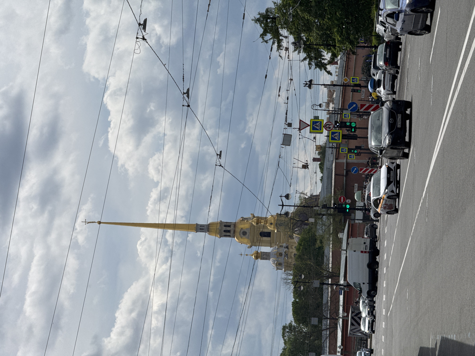 The image displays an urban street scene in Saint Petersburg, Russia, dominated by the prominent golden spire of the Peter and Paul Cathedral on the left. The photograph is taken from a street-level perspective, looking towards the cathedral, with a network of overhead wires for public transport crisscrossing the sky.
The Peter and Paul Cathedral features a tall, slender golden spire topped with a cross, characteristic of Russian Baroque architecture. Its main body is also golden, with several smaller domes and details visible. Green foliage of trees frames parts of the cathedral and lines the street on the right.
On the right side of the image, a multi-lane asphalt road stretches into the distance, filled with various cars and a white van, suggesting ongoing traffic. Many vehicles are parked or stopped in lanes. Green traffic lights indicate open traffic flow. A dense array of street signs is visible, including yellow diamond-shaped pedestrian crossing signs, a yellow triangular sign indicating a traffic light ahead, and a red circular "СТОП" (STOP) sign. One circular sign appears to indicate a speed limit, possibly "60". Red-brick buildings with windows line the right side of the street, behind the cars.
The sky is partly cloudy with patches of brighter blue, indicating daylight conditions, likely during the late morning or afternoon. Several Russian license plates are visible on the cars, confirming the location with region codes like "178" and "98" (Saint Petersburg). The overall scene captures a busy city street with a historic landmark under a bright, cloudy sky.