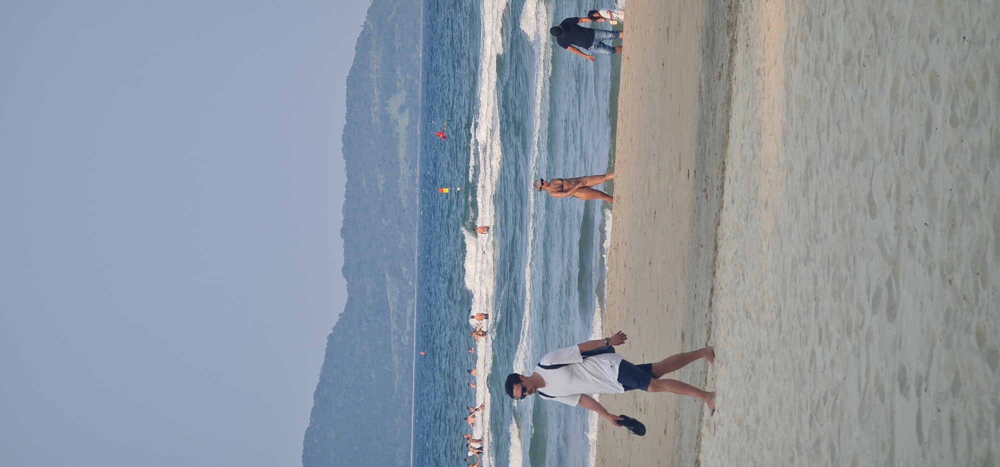 This image captures a lively beach scene in Đà Nẵng, Vietnam, likely during the late afternoon or early morning due to the soft, diffused lighting and light blue, hazy sky. The scene is dominated by the ocean on the left with gentle breaking waves, a wide expanse of light-colored sand on the right, and a backdrop of large, tree-covered mountains under the clear sky.
In the foreground, on the sandy beach, a man wearing a white t-shirt, dark shorts, and sunglasses walks barefoot, carrying a pair of black flip-flops in his hand. He is facing the ocean, heading towards the water.
Closer to the water's edge, a woman in a dark bikini and a hat stands in the shallow waves, looking out to sea. Further down the beach, another man in a dark t-shirt and jeans is partially submerged in the water, accompanying a small child. The ocean is dotted with numerous other people, some wading, some swimming, and others standing in the surf, enjoying the refreshing water.
In the distance within the ocean, a red flag and a yellow-and-red flag are visible, likely marking designated swimming areas or indicating beach conditions. The weather appears clear and warm, conducive to beach activities. The overall mood is relaxed and recreational, with many individuals enjoying the natural beauty of the coastline. There is no visible text in the image.