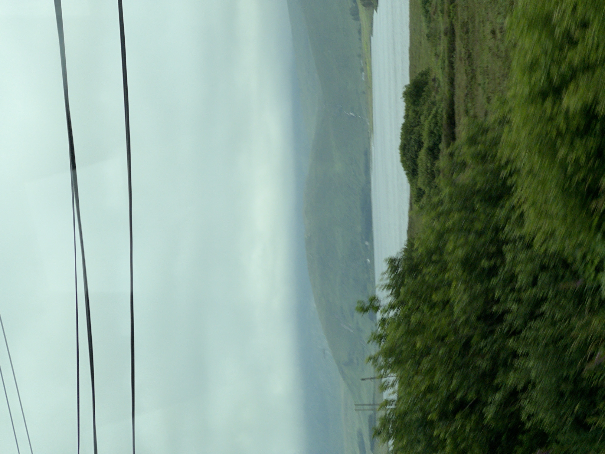 The image shows a landscape scene with a body of water in the middle ground, bordered by rolling green hills under a cloudy sky. In the foreground, there is dense, blurred green foliage, suggesting movement or a close-up perspective through vegetation. Overhead, several dark, thin wires are visible, stretching across the frame. The distant hills appear soft and hazy due to atmospheric conditions. The overall impression is one of a natural, possibly remote, environment, with the wires indicating human presence or infrastructure within the landscape. The scene is likely a view from a moving vehicle, given the blurriness of the foreground foliage. The location is identified as Meenacuing, Ireland.