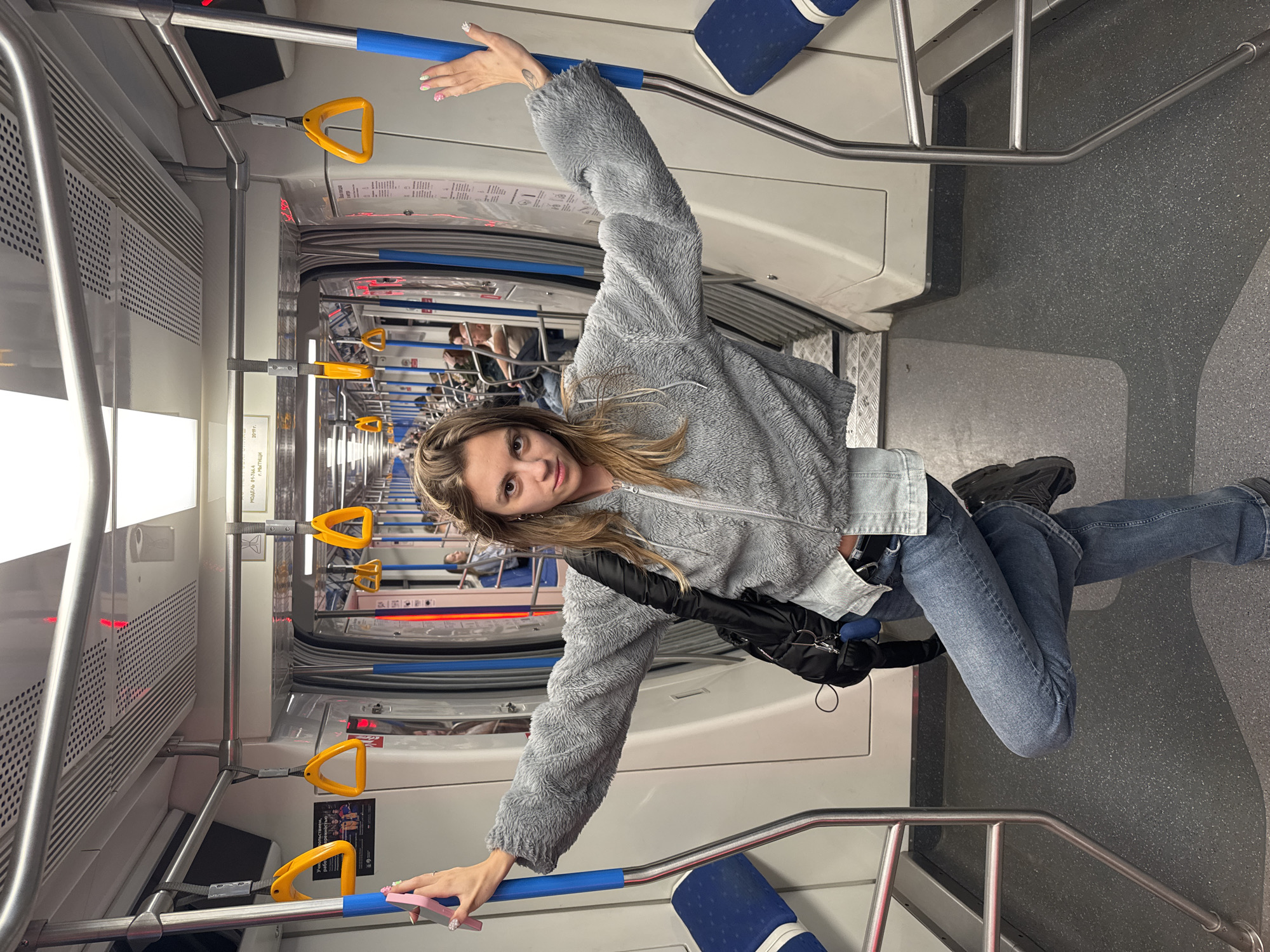 A woman stands with her arms outstretched inside a Moscow Metro train car, leaning against a pole with her right hand and holding a pink smartphone in her left. She is wearing a grey fuzzy jacket, a denim shirt, and blue jeans. Her legs are crossed, with her left leg bent at the knee. The train car is mostly empty, with a few other passengers visible in the background. The interior of the train is modern and clean, with white walls, blue seats, and yellow handrails. The floor is dark grey with a speckled pattern. The lighting is bright and even.