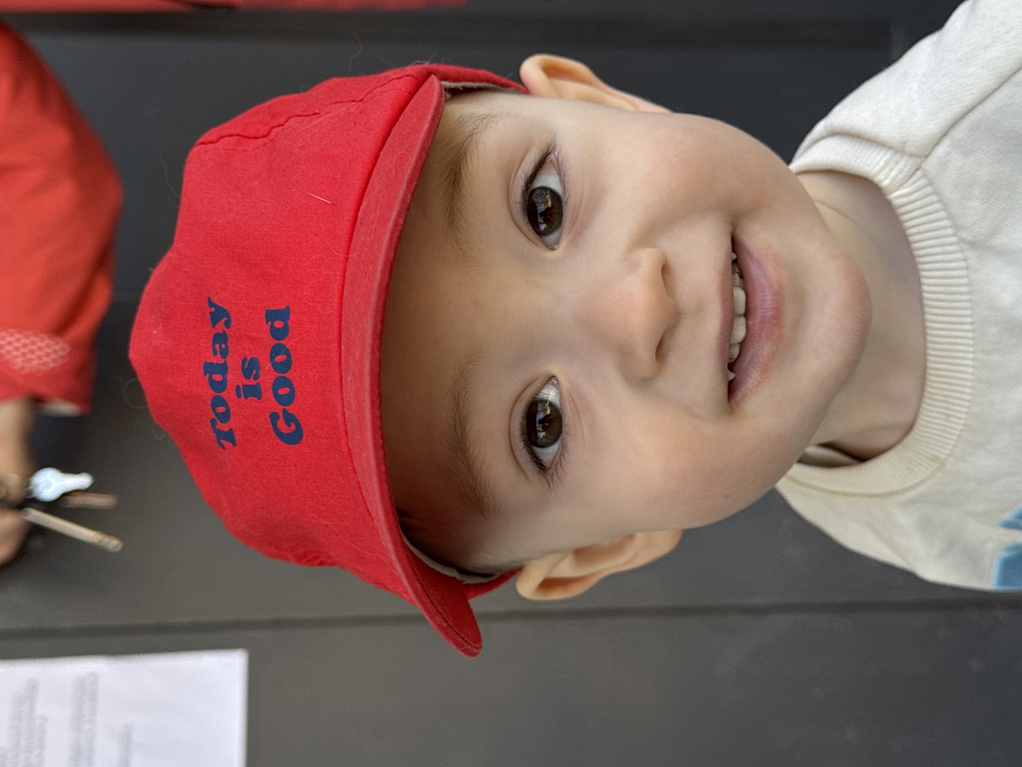 A young child with fair skin is smiling while wearing a red baseball cap with the words "Today is Good" in blue lettering. The child is also wearing a light-colored, long-sleeved shirt. The background appears to be a gray surface, possibly a table. Part of another person wearing a red outfit is visible in the corner. There is no information in the image to confirm the location is Izhevsk, Russia.