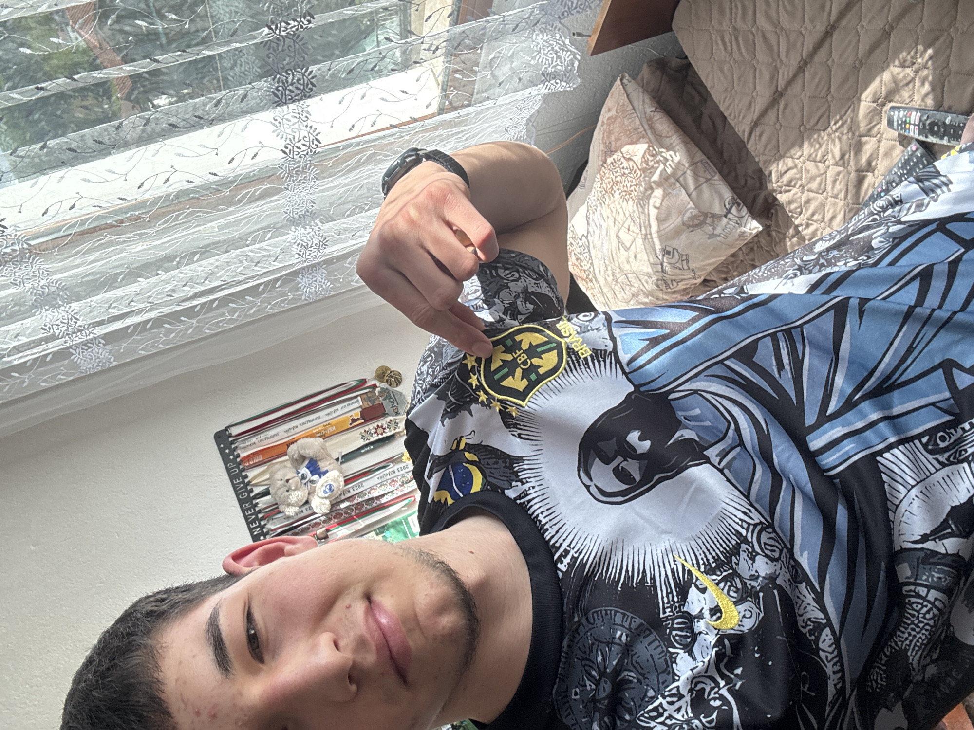 An indoor, bright, daytime selfie taken in Zlatograd, Bulgaria shows a young man from the chest up, looking towards the camera with a slight smile. The image is oriented sideways. He has short dark hair and light facial hair.
The main subject is wearing a multi-colored t-shirt with intricate patterns in black, white, and blue. He is pointing with his right hand to a yellow and green emblem on the shirt's sleeve, which displays "CBF" and partially visible "BRASIL," indicating the Brazil Football Confederation. A golden Nike swoosh is also visible on the shirt's design. A dark-colored watch is on his left wrist.
In the background to the left, bright sunlight streams through a window covered by patterned white sheer curtains or blinds, casting shadows. To the right, part of a bed or sofa is visible with a quilted brown blanket and a patterned pillow. A TV remote control rests on the blanket. Behind the man's head, there's a shelf or rack filled with books or binders, alongside two small plush toys.
Visible text from the background items includes: "NEVER GIVE UP", "GOLD D", "2023 KJULTRA", "2023 K", and "$3300". The overall scene suggests a relaxed home environment.