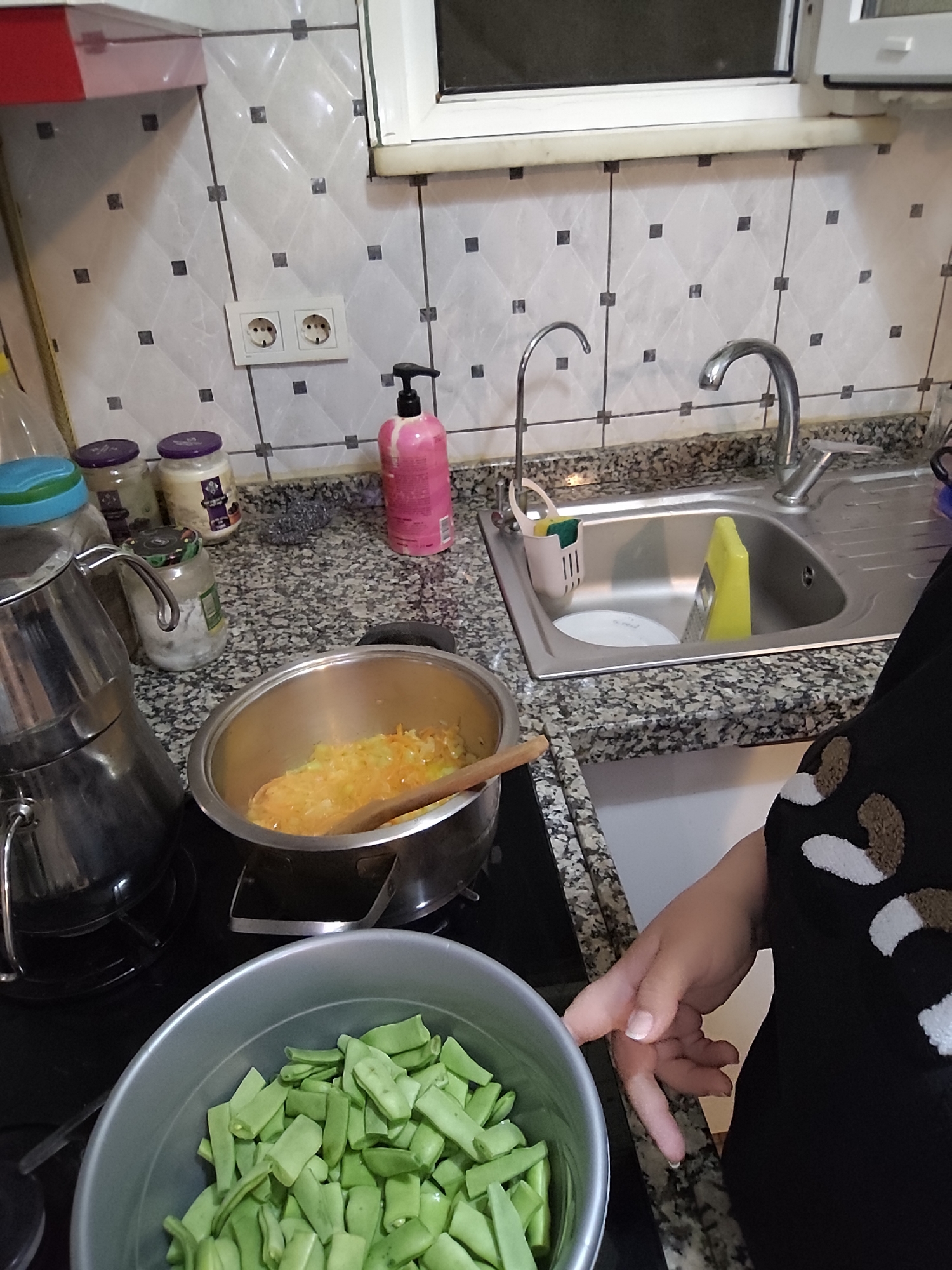 A person is in a kitchen in Sarıyer, Turkey, preparing a meal. A pot of cooked vegetables sits on a stovetop next to a kettle. A bowl filled with chopped green beans is in the foreground. A kitchen sink with a faucet is on the counter. Jars and a soap dispenser are also on the counter. The background features wall tiles and a window.