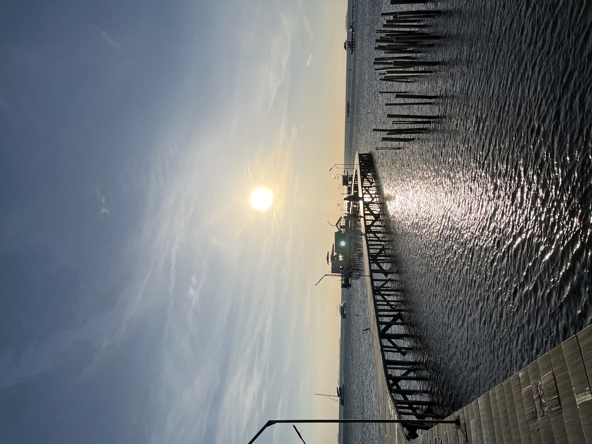 This is a wide shot taken at sea level, featuring a wooden pier that curves out into the water, leading to small structures. The sun is low in the sky, casting a warm glow and creating a shimmering reflection on the rippling water. The sky is a clear blue with wispy clouds. In the distance, there are several boats on the water and landmasses on the horizon. The scene appears to be located in Karimunjawa, Indonesia.