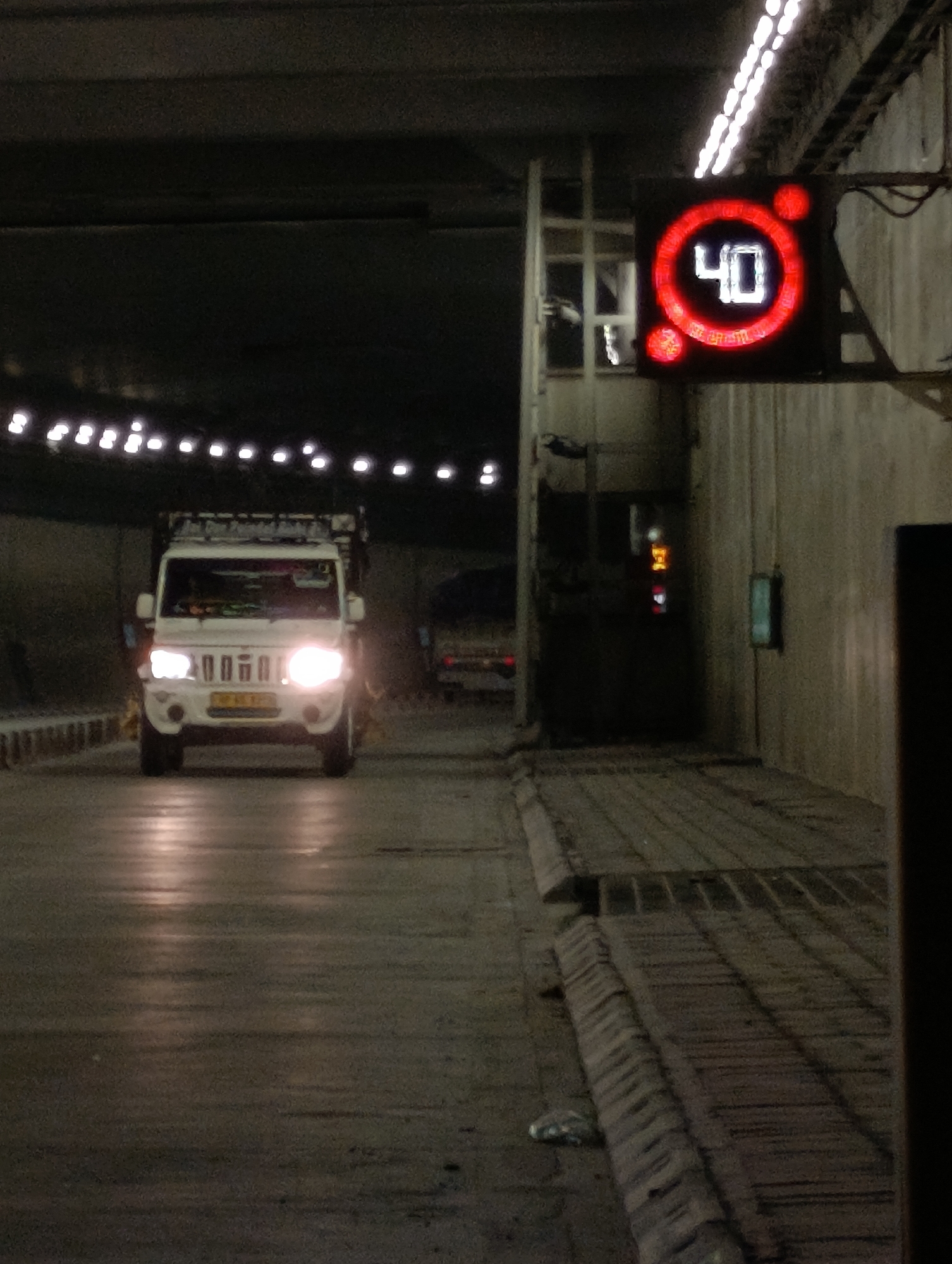 In an underground tunnel, a white pickup truck with its headlights on drives forward. A digital speed limit sign displaying "40" in red is visible in the upper right corner of the image. The tunnel is lit by overhead lights, and the road surface appears wet, reflecting the lights.