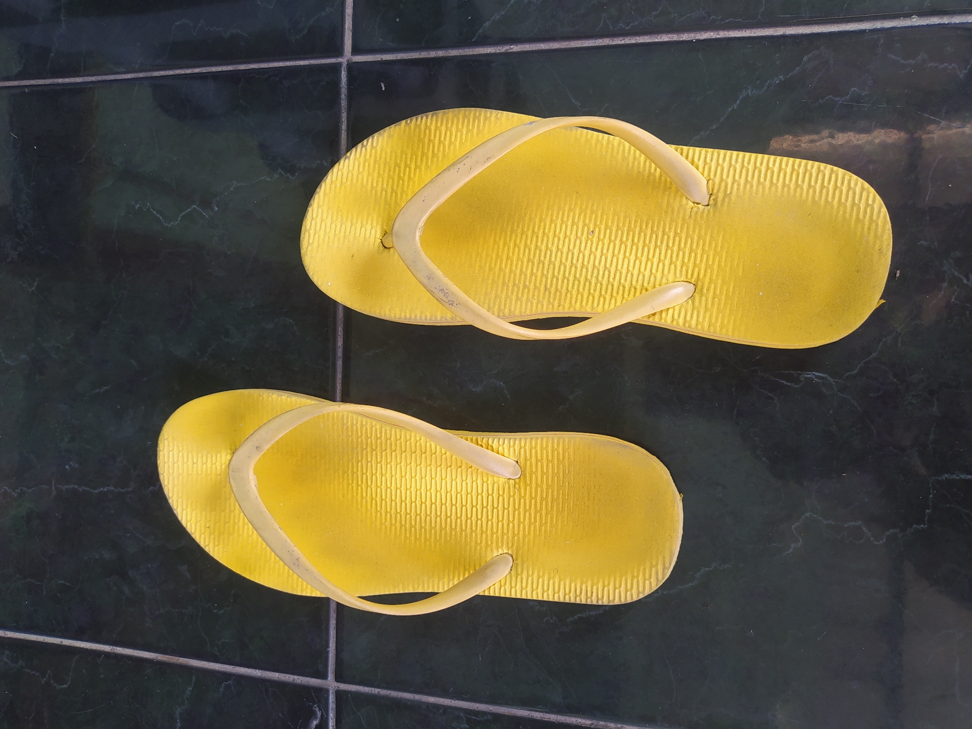 Two bright yellow flip-flops are displayed on a dark green tiled floor. The tiles have a subtle marble-like pattern. The flip-flops are positioned side-by-side, with the left flip-flop slightly angled towards the top left and the right flip-flop angled towards the bottom right. Both flip-flops are oriented with the toe pointing upwards in the frame. The surface of the flip-flops exhibits a textured pattern, and there are minor signs of wear, such as slight scuffing and a few specks of dust. The lighting suggests an indoor environment, casting faint shadows. The location is identified as Sroyo, Indonesia.