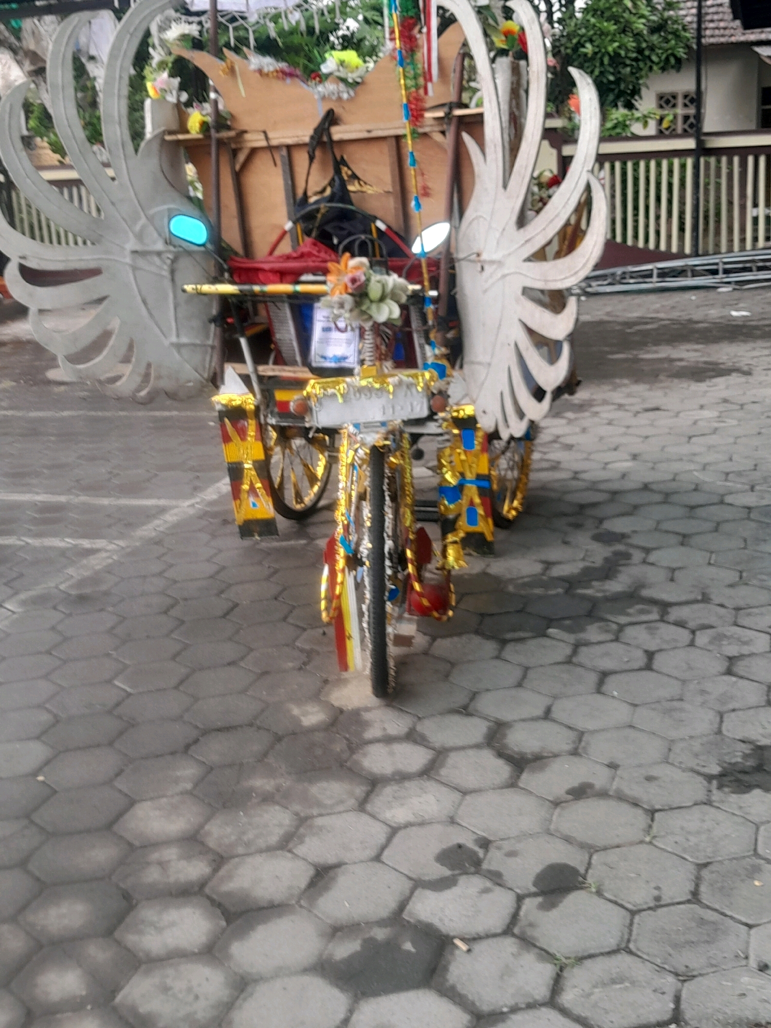 An outdoor, eye-level, full shot shows a heavily decorated, three-wheeled vehicle parked on a hexagonal-tiled surface. The vehicle is adorned with large, white, wing-like structures on either side of its rear, and features intricate gold and blue decorative elements along its body and wheels. Flowers and other decorative items are attached to the vehicle. The background reveals a building with white walls and a tiled roof, bordered by a wooden fence. Lush green trees are visible behind the fence. The lighting suggests it is daytime.