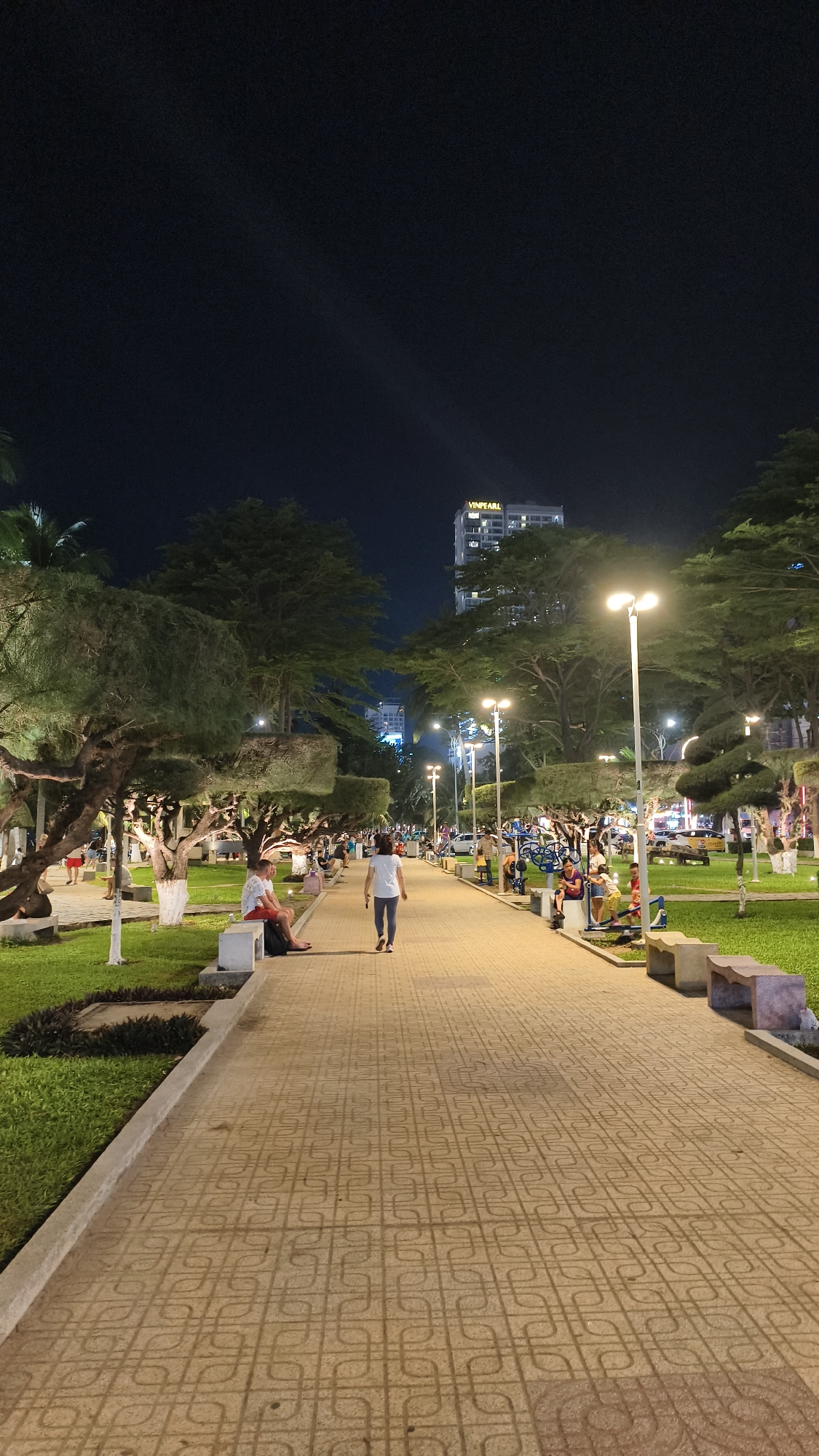 This is a vertical, eye-level, nighttime, outdoor, full shot of a pedestrian walkway in Nha Trang Ward, Vietnam. The walkway is paved with interlocking square tiles, and it is lined with trees on both sides, with grass areas between the trees and the walkway. Several streetlights illuminate the path.
In the foreground, on the left side of the walkway, a man wearing red shorts and a white t-shirt is sitting on a stone bench, facing right. To his right, a woman in a white t-shirt and dark pants is walking away from the camera down the center of the walkway. Further down the walkway, on the right side, there are several people and exercise equipment. A tall, modern building with the word "VINPEARL" illuminated in yellow is visible in the background, behind the trees. The sky is dark, indicating it is nighttime.