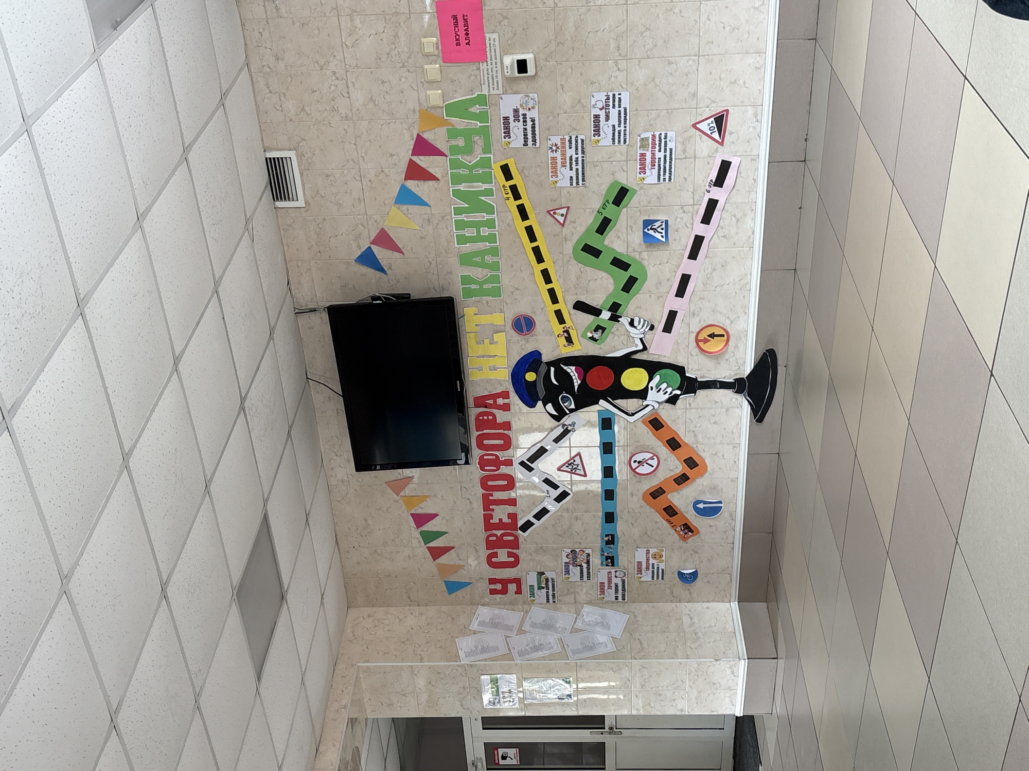 The image depicts an indoor hallway or common area, likely within a school or children's summer camp in Birobidzhan, Russia. The walls are covered in light-colored tiles, and the ceiling is a standard drop-tile design with some fluorescent lighting panels.
The primary subject is a large, colorful educational display mounted on one wall. This display features a stylized figure resembling a traffic light with a head, arms, and legs made from cutouts resembling road segments. The main title across the top reads "У СВЕТОФОРА НЕЙ КАНИКЧА," roughly translating to "At the Traffic Light (on) Holidays," suggesting a themed educational activity or camp program focused on road safety during vacations.
Various Russian road signs are incorporated into the display, illustrating different traffic regulations such as pedestrian crossings, no entry, speed limits, and turns. Several smaller signs, each titled "ЗАКОН" (LAW/RULE), are interspersed throughout the display. These rules cover a range of topics, including mutual help, unity, precision, creativity, promoting a healthy lifestyle ("ЗОЖ - береги своё здоровье!"), respect ("если хочешь, чтобы уважали тебя, относись с уважением к другим"), cleanliness ("соблюдай гигиену, содержи вещи в чистоте и порядке!"), and a specific rule for a camp setting: "запрещается выходить за территорию лагеря без предупреждения!" (it is forbidden to leave the camp territory without warning!). Some road segments are also numbered, like "4 ОТP," "5 ОТP," and "6 ОТP," possibly indicating different groups or activity stages.
A black flat-screen television is mounted on the wall to the left of the main display. A colorful banner made of triangular flags hangs nearby. Other notable details include a pink sign labeled "ВКУСНЫЙ АЛФАВИТ" (TASTY ALPHABET) and several smaller white papers taped to the wall below the main display. The scene suggests an educational and informative environment designed for children, possibly as part of a summer camp or a school's road safety initiative.