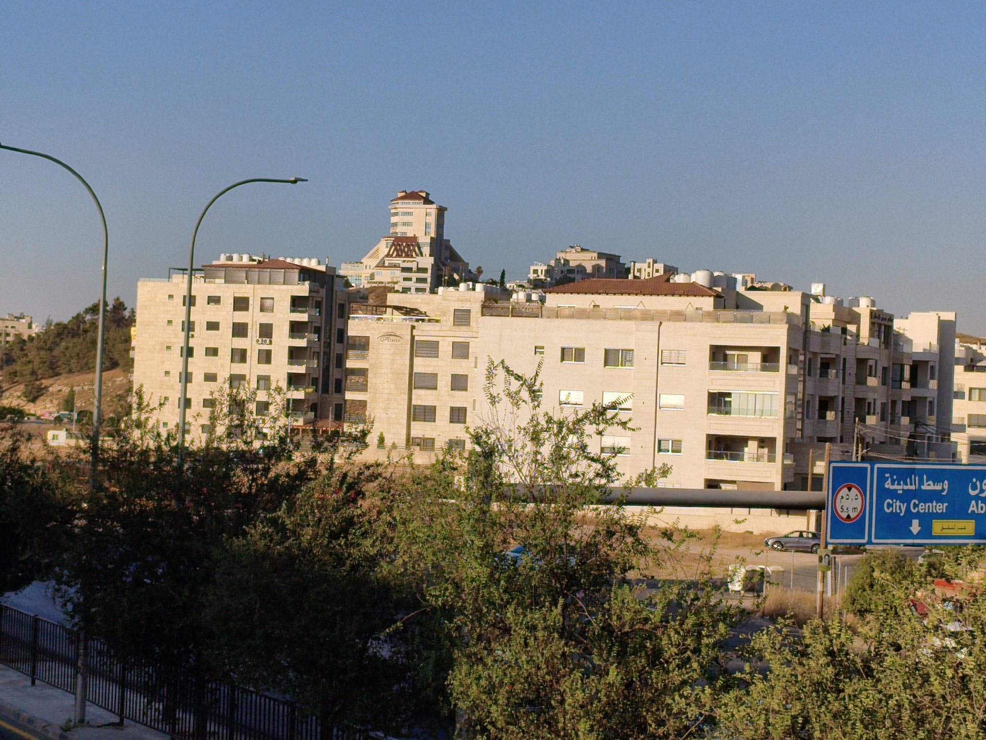 The image depicts a cityscape in Amman, Jordan, with multiple buildings and a clear blue sky. In the foreground, lush green trees partially obscure a black metal fence and a walkway. Beyond the trees, several modern apartment buildings are visible, constructed with light-colored stone. The buildings exhibit a consistent architectural style with numerous rectangular windows and balconies.
Further in the background, a distinctive building with a stepped, pyramidal roof structure stands out, suggesting it might be a landmark or a larger complex. To the right, a blue road sign is prominently displayed, featuring Arabic text and English text that reads "City Center" and "Ab". A speed limit sign with "5.5 m" is also visible on the blue sign. A silver car is parked on the side of the road near the sign. The overall impression is one of a developing urban environment under bright sunlight.