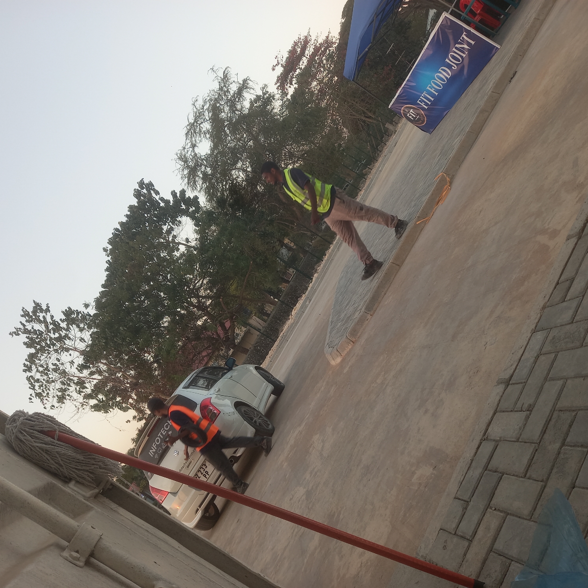 In Bububu, Tanzania, two men in high-visibility vests are visible on a paved area near a white Suzuki Swift. One man, wearing a fluorescent orange vest over a dark shirt and dark pants, is positioned near the rear of the car, appearing to interact with its rear bumper. The second man, in a fluorescent yellow-green vest over a dark shirt and light brown pants, stands further away near the roadside, facing the car. The car has a license plate that reads "Z 233 PP" and a sticker on its rear window says "INFOTECH SOLUTION.CO". A red mop is leaning against a light-colored wall to the left of the frame. In the background, trees with dense foliage are prominent, and a blue tent with a banner reading "FIT FOOD JOINT" is visible. The ground is a mix of paved concrete and interlocking pavers.