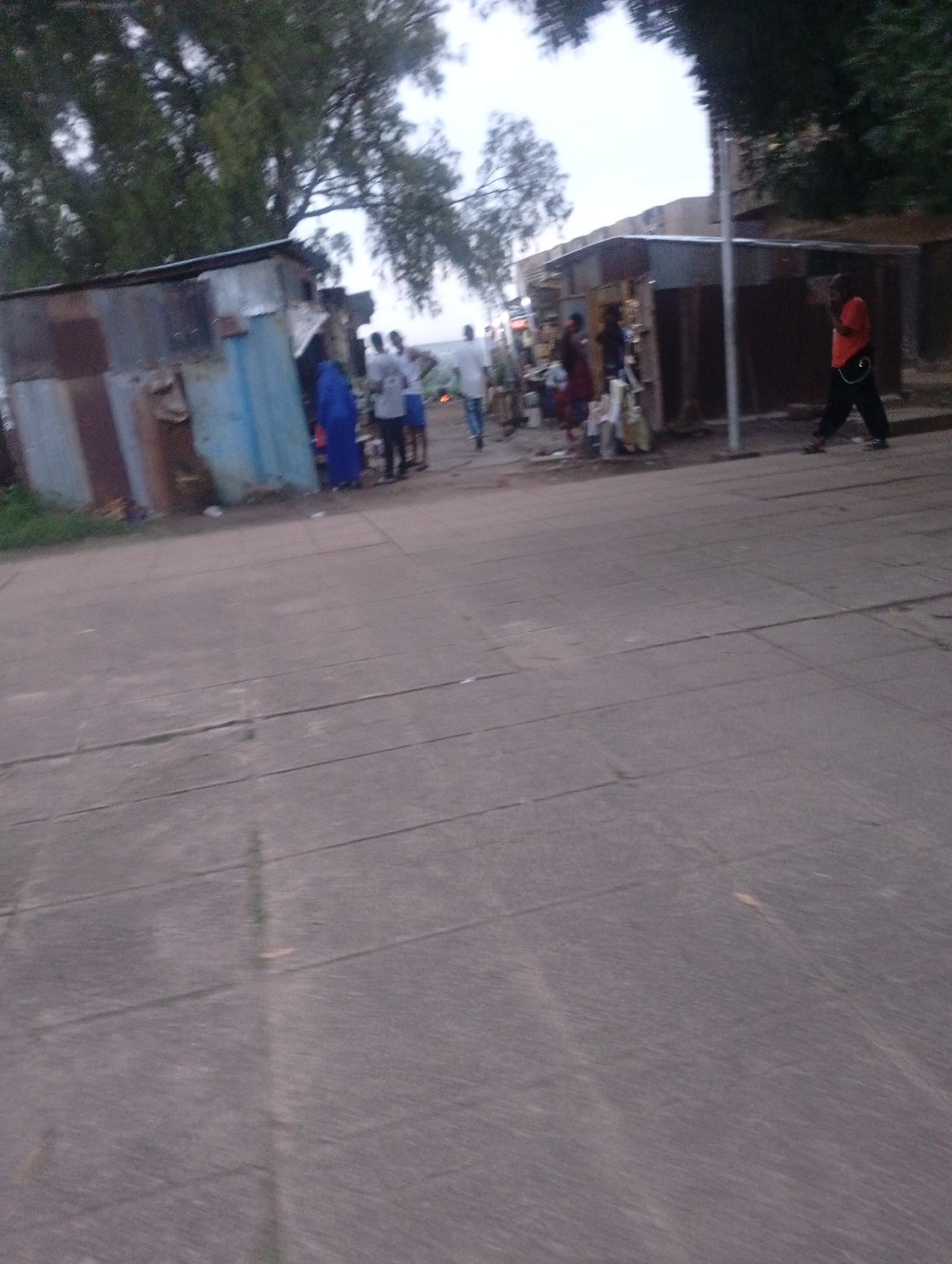 The image depicts a street scene in Jos, Nigeria. In the foreground, a paved area, possibly a sidewalk or street, shows signs of wear and tear with visible cracks and unevenness. In the background, several makeshift structures, likely stalls or informal shops, are visible. These structures appear to be constructed from corrugated metal and possibly wood. Several people are present around these structures. One individual, wearing an orange shirt and dark pants, is walking away from the camera on the right side of the image. Other individuals are gathered near the stalls, with some appearing to be interacting or engaged in activities. The overall environment suggests a bustling, informal marketplace or a residential area with commercial activity. The lighting indicates it might be late afternoon or early evening.
