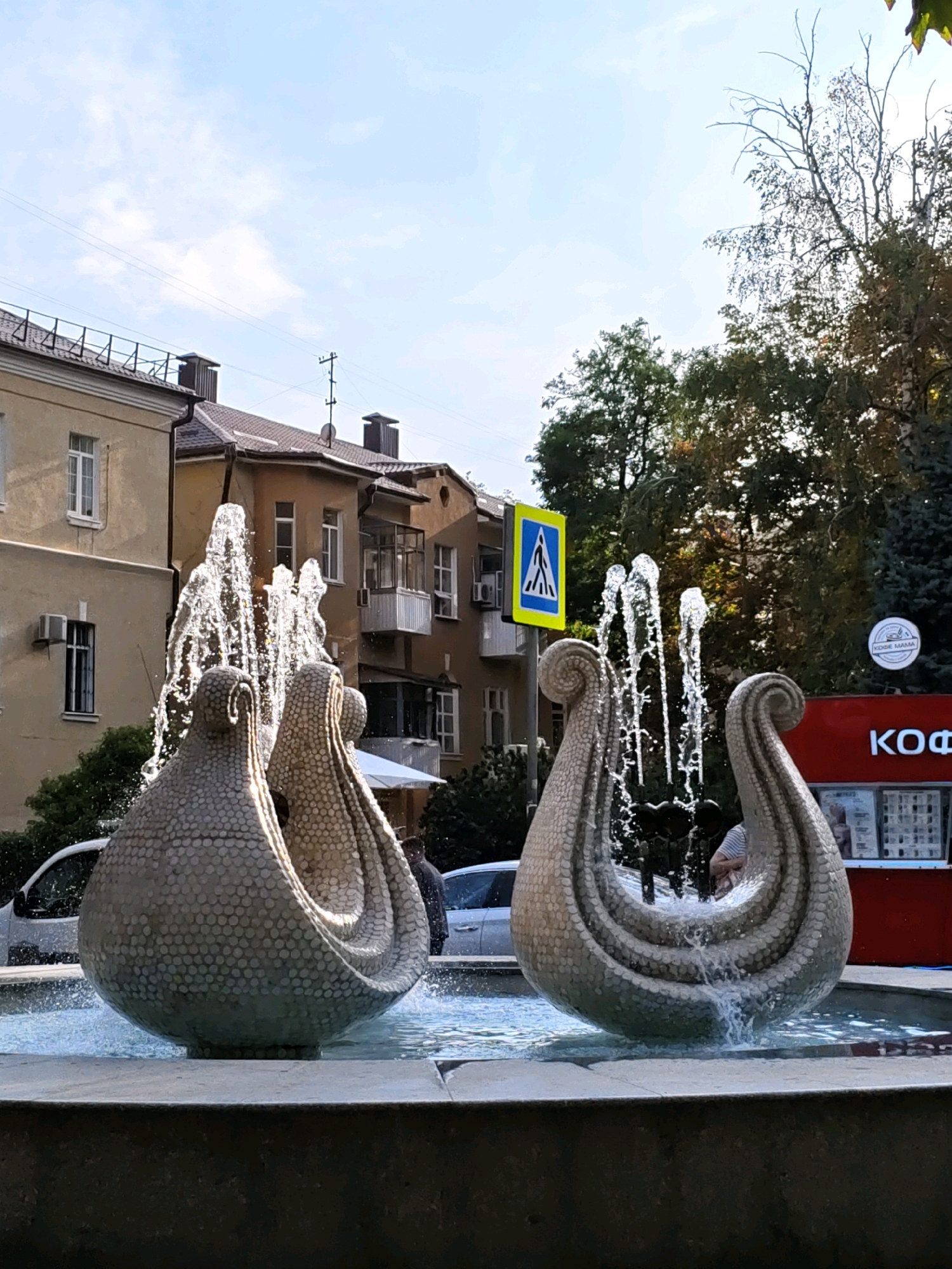 This is a medium-angle, eye-level shot of a public fountain featuring two large, ornate sculptures that resemble abstract, stylized seed pods or shells. The sculptures are made of a light-colored material covered in small, circular indentations, giving them a textured, almost scaly appearance. Water sprays upward from the center of each sculpture and cascades down their sides, creating a dynamic visual effect. The fountain is situated in a paved area, with a concrete edge in the foreground.
Behind the fountain, the scene depicts an urban environment. On the left, there are multi-story buildings with a light yellow facade, featuring windows and balconies. Air conditioning units are visible on the walls. In the center and right, more buildings with brown and cream facades are visible, some with balconies. A red kiosk or small building with the Cyrillic letters "КОФ" (likely part of a word related to coffee) is on the right side of the frame.
A yellow pedestrian crossing sign with a white silhouette of a person walking is visible behind the fountain on the left. Trees with green and autumn-colored foliage are present in the background, suggesting it is either late summer or early autumn. The sky is partly cloudy with blue patches. Two white cars are parked or driving near the fountain. A few individuals are partially visible in the background.
This fountain is located on Bolshaya Sadovaya Street in Rostov-on-Don, Russia, and is a well-known landmark.