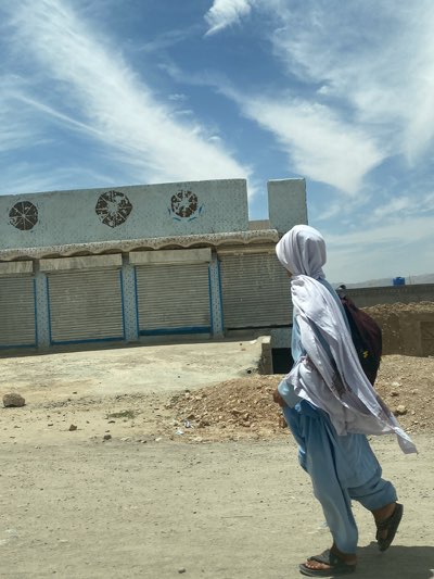 A person wearing traditional clothing and carrying a backpack walks along a street in Sard-E 'olya, Afghanistan. The person is dressed in light blue clothing with a white headscarf, and is wearing sandals. They are walking towards the right side of the image. Behind the person is a building with closed metal shutters and faded circular designs on the upper part of the facade. The surrounding environment appears arid and dusty. The sky is partly cloudy with streaks of white clouds.