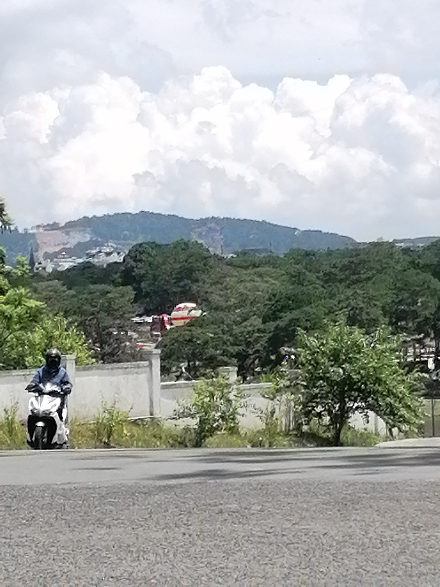 A person wearing a dark helmet and blue jacket rides a white scooter on a paved road in Phường Xuân Hương - Đà Lạt, Vietnam. The scooter is partially obscured by the rider's body and is positioned in the lower left quadrant of the image. In the background, a low, white wall separates the road from a treeline. Beyond the wall, dense green trees are visible, with a white and red object, possibly an emergency vehicle, partially hidden within the foliage. Further in the background, a hillside is visible with signs of construction or development, and a cloudy sky with large white clouds dominates the upper portion of the frame.