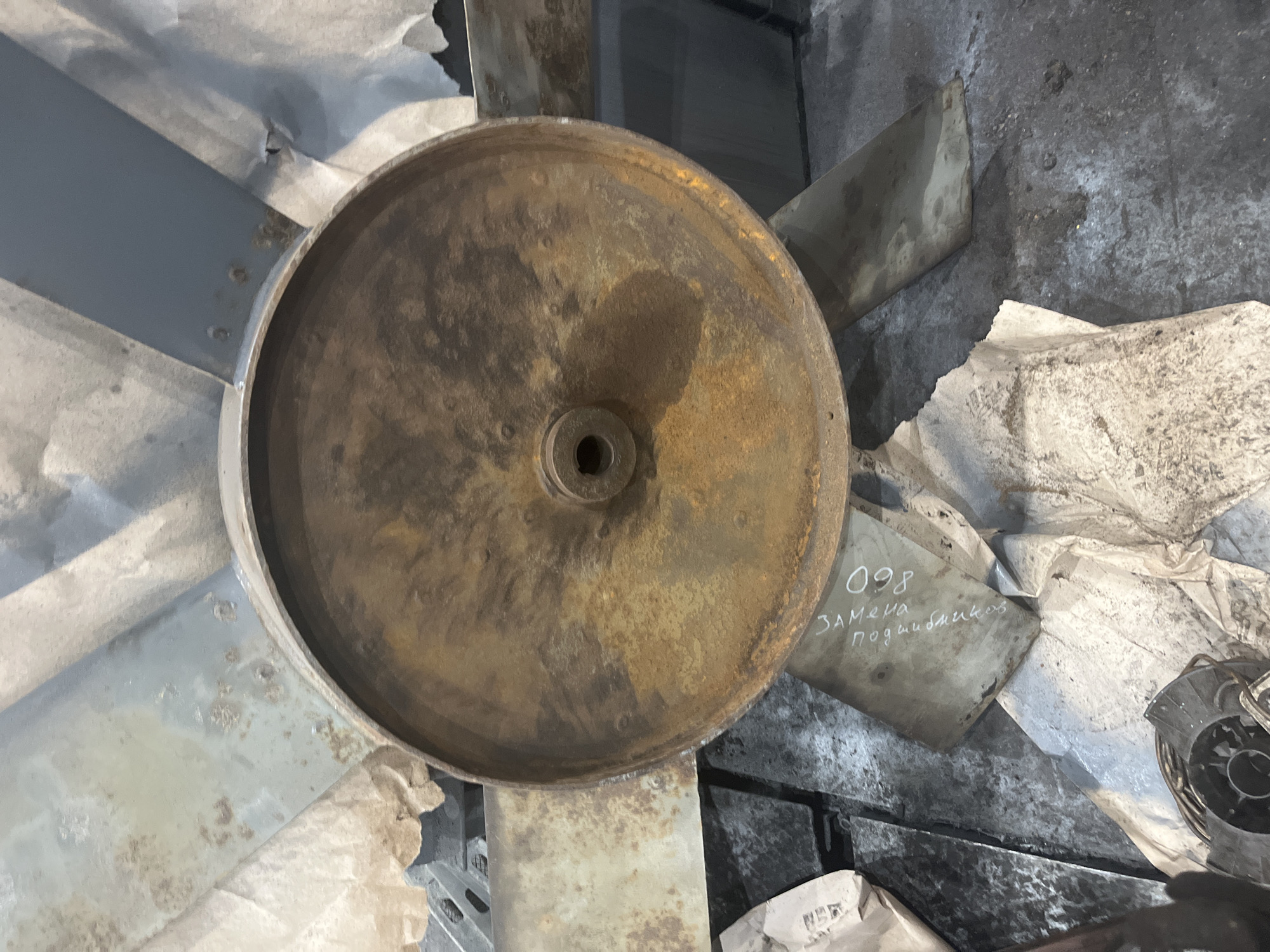 The image shows a corroded metal fan assembly lying on a surface littered with debris, including what appears to be crumpled paper or cloth and discarded metal parts. The fan itself is rusty and displays signs of wear and tear. One of the blades has handwritten text in Russian, reading "098 ЗАМЕНА ПОДШИПНИКОВ," which translates to "098 Replacement Bearings." The overall setting seems to be an industrial or storage area, and based on the inscription in Russian, it's located in Russia, likely in the city of Chaikovsky.