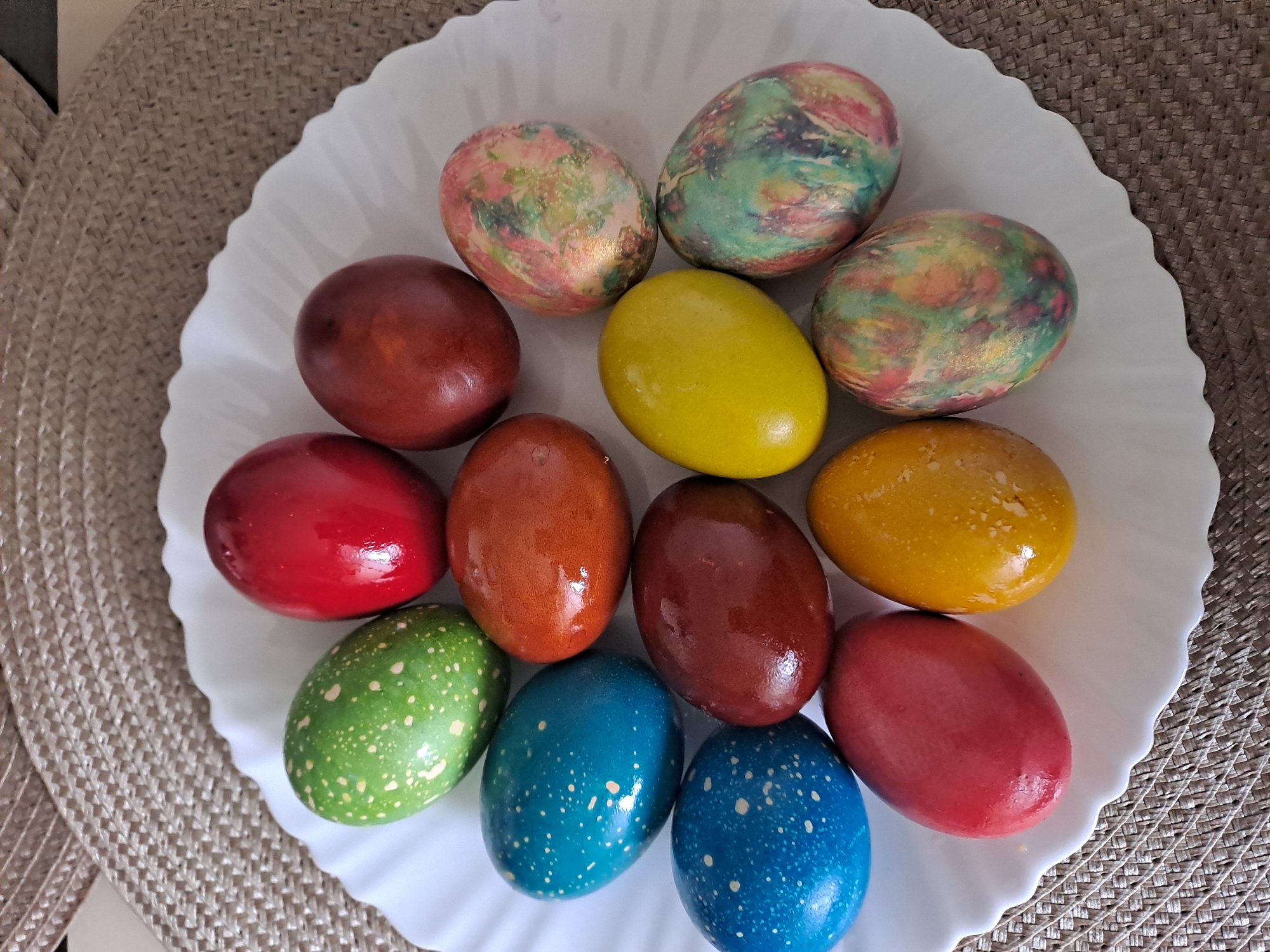 A white, ruffled-edged plate sits on a textured, beige placemat. Arranged on the plate are twelve Easter eggs of various colors and designs. Starting from the top left and moving clockwise, there is an egg with a mottled pattern of pink, green, and gold; an egg with a mottled pattern of green, blue, pink, and gold; an egg with a mottled pattern of green, gold, and pink; a solid yellow egg; a solid golden-yellow egg; a solid red egg; two solid reddish-brown eggs; a blue egg with white speckles; a green egg with white speckles; a solid red egg; and a solid orange egg. The image was likely taken indoors. There is no indication of the location of the image being in Elektrostal, Russia, or of any people, subjects, actions or interactions.
