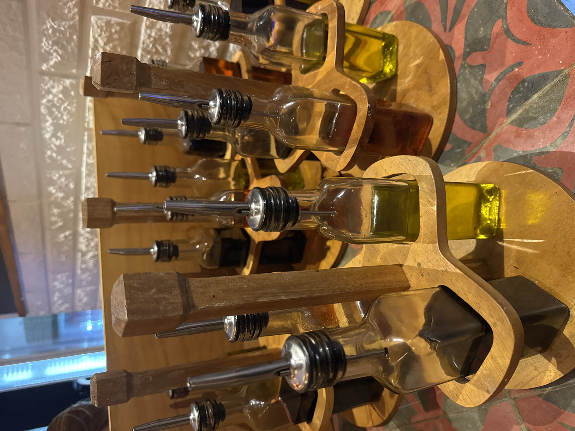 This image captures a display of numerous glass bottles, each fitted with a metal pour spout, arranged on tiered wooden racks. The bottles appear to hold various culinary liquids, predominantly golden yellow oil, likely olive oil, and some darker liquids, possibly vinegars or other flavored oils. They are held within individual, shaped wooden cradles that are part of larger wooden stands.
The display is vertically oriented, with multiple rows of bottles filling the frame. The wooden stands are crafted from light-colored wood, exhibiting a smooth, finished surface. Each bottle is clear glass, allowing the liquid inside to be visible. The metal spouts are shiny and reflect some light.
In the background, a light-colored wall with a rough, textured, brick-like pattern is visible on the left side of the image. On the right and bottom, part of a floor covered with decorative tiles is visible. These tiles feature a distinct pattern of red, dark grey, and lighter grey organic shapes, resembling leaves or stylized floral motifs.
No people are present in the image, and no specific activity is taking place; the bottles are static and presented for display or use. The lighting suggests an indoor setting, possibly a restaurant, deli, or specialty food store, in Kuta Selatan, Indonesia. There is no visible text in the image.