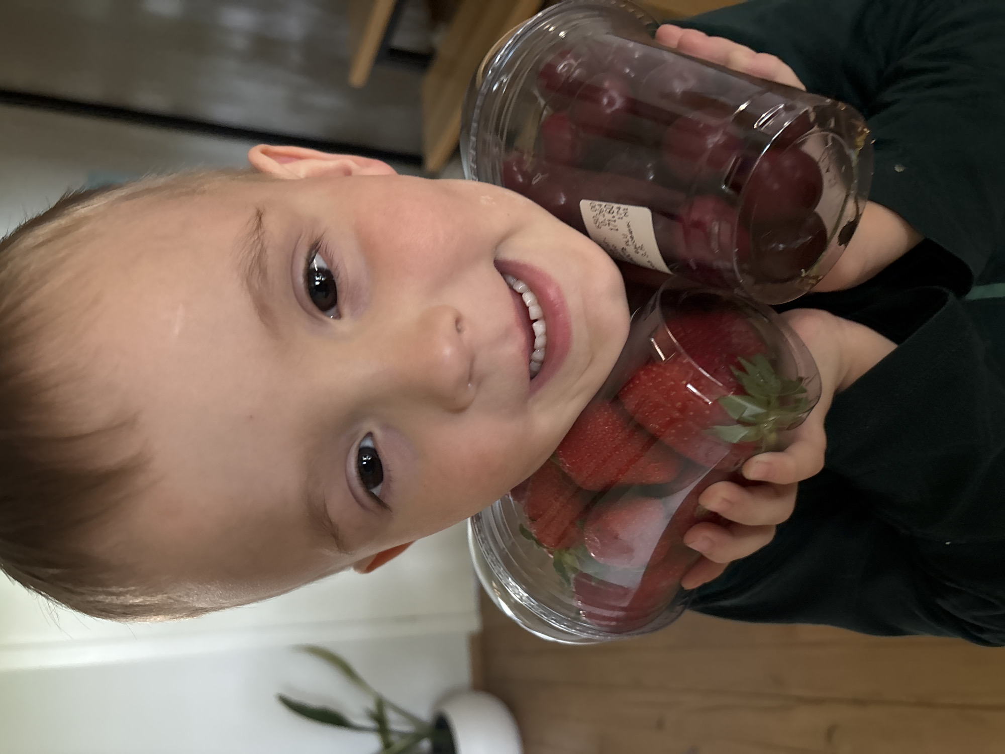A young child holds containers of strawberries and cherries. The child is fair-skinned with brown hair and eyes, and is smiling, revealing teeth. The child is wearing a dark green shirt. The strawberries are in a clear plastic container with green leaves visible. The cherries are also in a clear plastic container. The background includes a wooden floor, a potted plant with green leaves, and a glimpse of what appears to be a cabinet or shelving unit. The location is not discernible from the image. There is no verifiable evidence to support that this photo was taken in Chaikovsky, Russia.