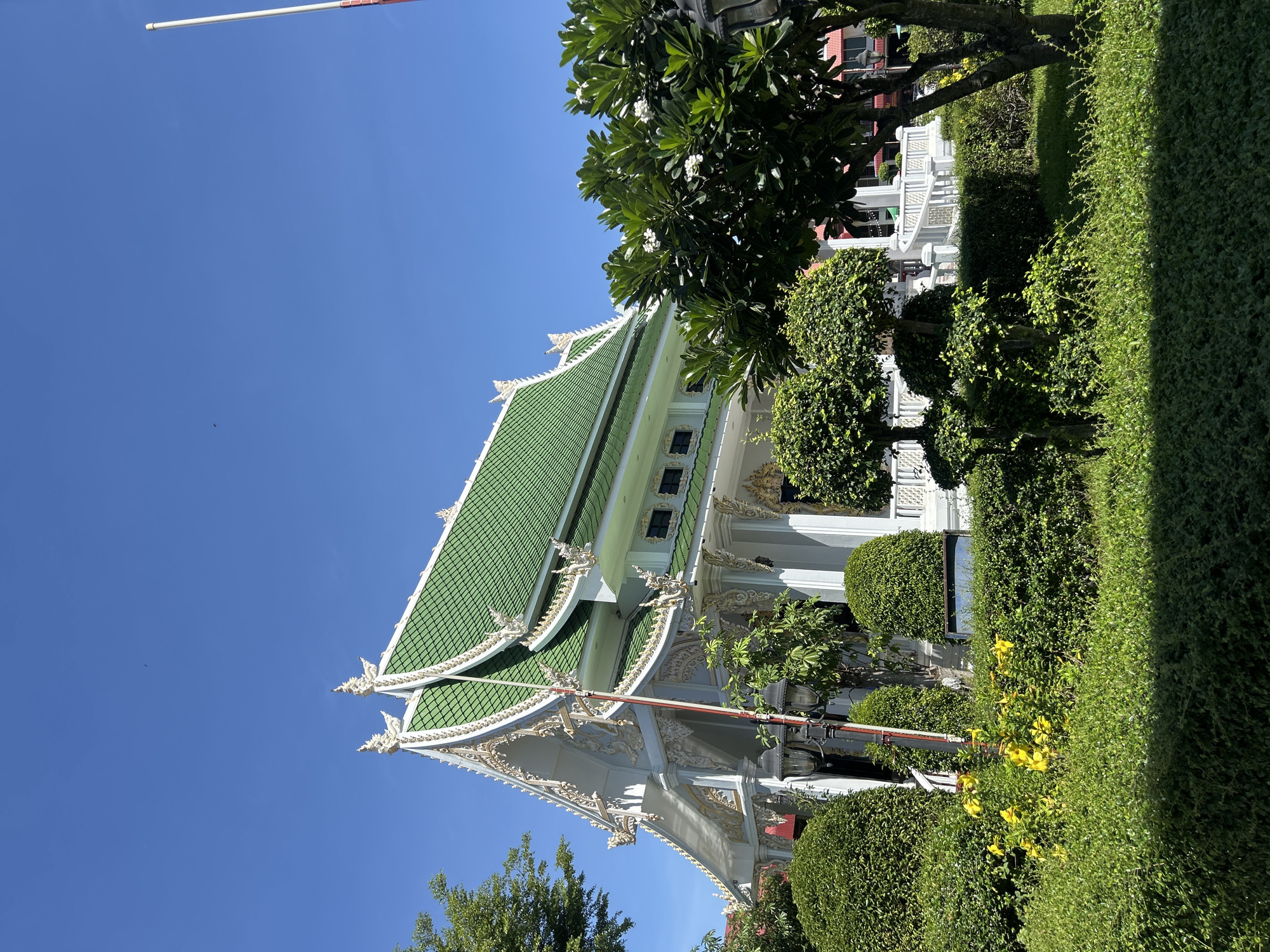 In Thai Ko Subdistrict, Thailand, a Buddhist temple stands with its distinctive green tiled roof adorned with ornate white and gold trim. The temple's facade is white with intricate decorative elements. Surrounding the temple is a meticulously landscaped garden featuring various shades of green bushes, manicured hedges, and a few scattered yellow flowers. A large tree with white blossoms is visible in the foreground on the right. The sky above is a clear, bright blue, indicating a sunny day. A tall flagpole stands on the right side of the frame, and a lamp post with two lamps is positioned in the garden. There are no people visible in the image.
