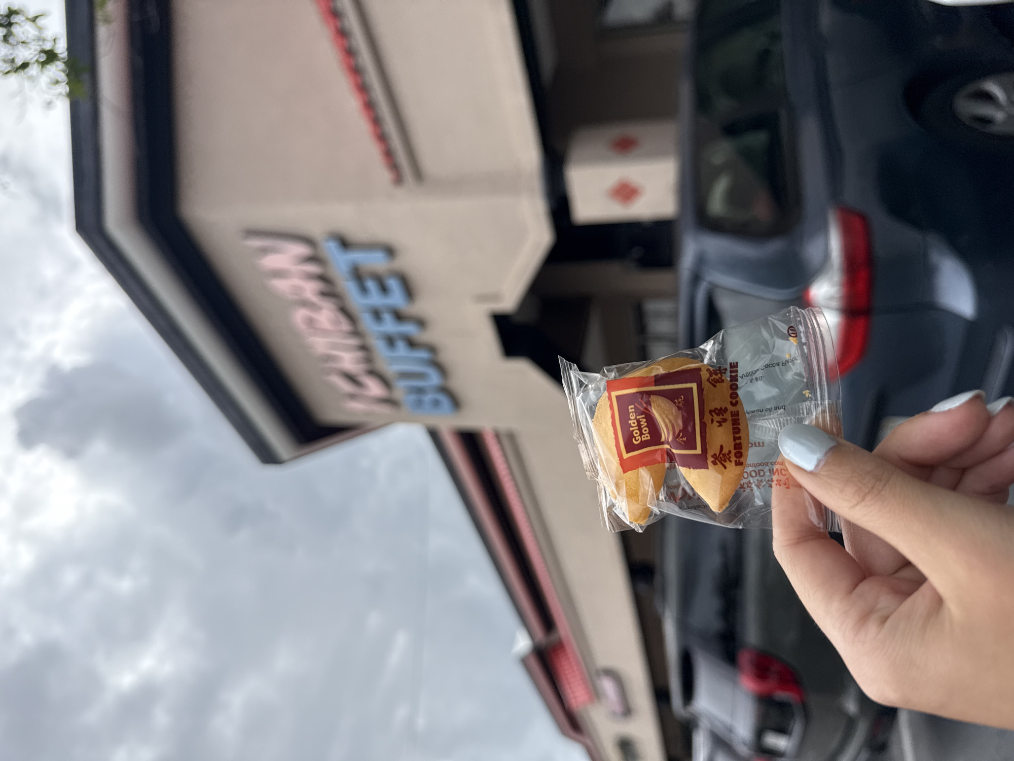 A person is holding a fortune cookie outside a building. The sign on the building reads "Ichiban Buffet". The fortune cookie's wrapper is labeled "Golden Bowl Fortune Cookie". The location appears to be in a commercial area, likely Kissimmee, United States of America. There are vehicles parked in the background, suggesting a parking lot. The sky is cloudy. The focus of the image is on the hand holding the fortune cookie, with the Ichiban Buffet sign acting as a background element.