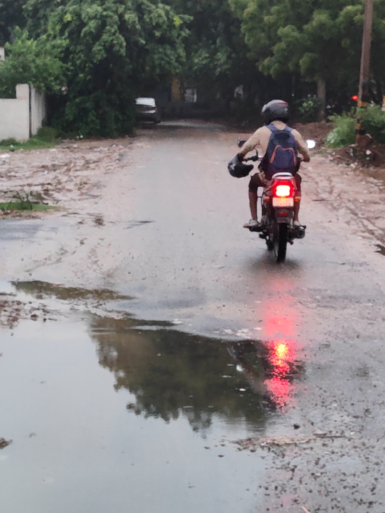 The image shows a person riding a motorcycle on a wet road in Gurgaon, India. The rider is wearing a helmet and a backpack, and the motorcycle's taillight is illuminated. The road appears to be in a somewhat neglected state, with puddles of water and debris scattered along the sides. Lush green trees line the background, suggesting a semi-urban or suburban environment. The sky is overcast, indicating that it might have recently rained or is about to rain. The overall scene conveys a sense of everyday life in a rainy climate, with the rider navigating the wet conditions.