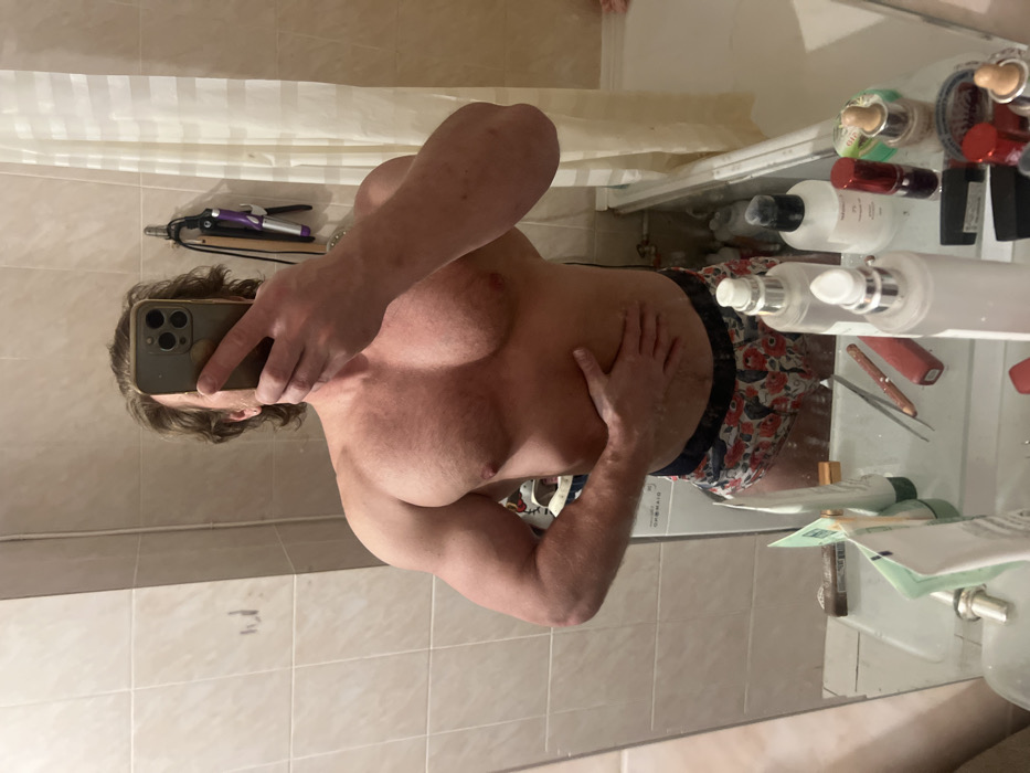 A shirtless individual is taking a mirror selfie in a bathroom. They have fair skin and are holding an iPhone. Their left hand is placed on their midsection. Floral patterned bottoms are visible. Various toiletries clutter the bathroom counter, including bottles and containers. A shower curtain hangs in the background. The bathroom has tile walls.