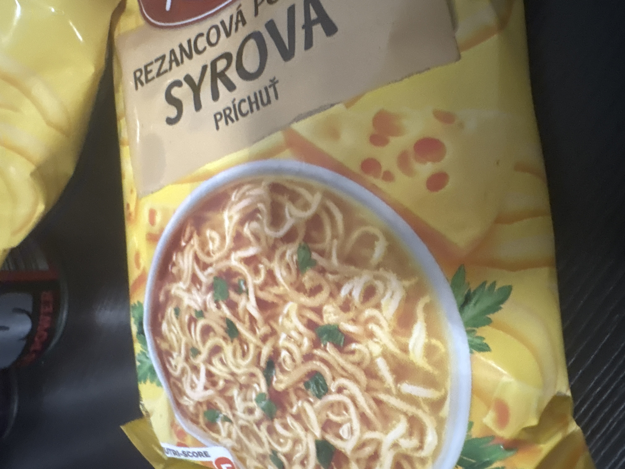 The image shows a close-up of a yellow package of instant noodles, with the text "REZANCOVA SYROVA PRICHUT" visible. A bowl of noodles is depicted on the left side of the package, garnished with parsley. The package itself has an image of cheese slices. The background is dark and appears to be a table surface. The lighting is dim, and the image is slightly blurry, suggesting it was taken in low light conditions with a handheld device. No specific location within a city is identifiable.