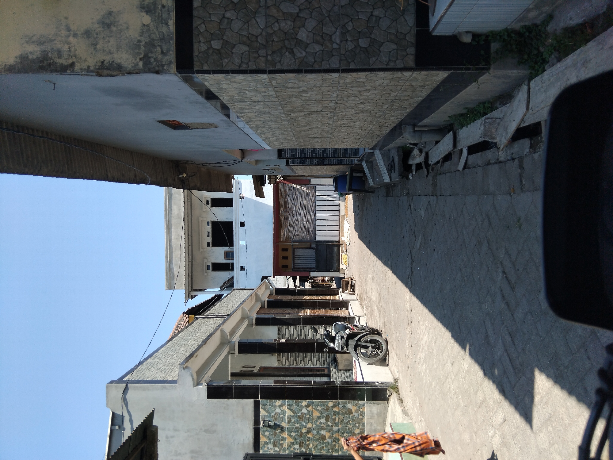 A street in Kepuh Kiriman, Indonesia, is captured on a sunny day. The narrow street is paved with interlocking bricks and is flanked by buildings on both sides. On the left, a two-story building has a facade with grey stone-like tiles and black pillars. A motorcycle is parked on the side of the road in front of this building. Further down the street on the left, a person wearing a patterned sarong is walking.
On the right, a building features a textured stone-patterned facade with black accents. A row of wooden planks is placed along the base of this building, partially covering a drainage ditch. In the background, a white building with multiple windows is visible, and further beyond, a blue barrel sits next to a building with a corrugated metal roof. The sky is clear and blue.