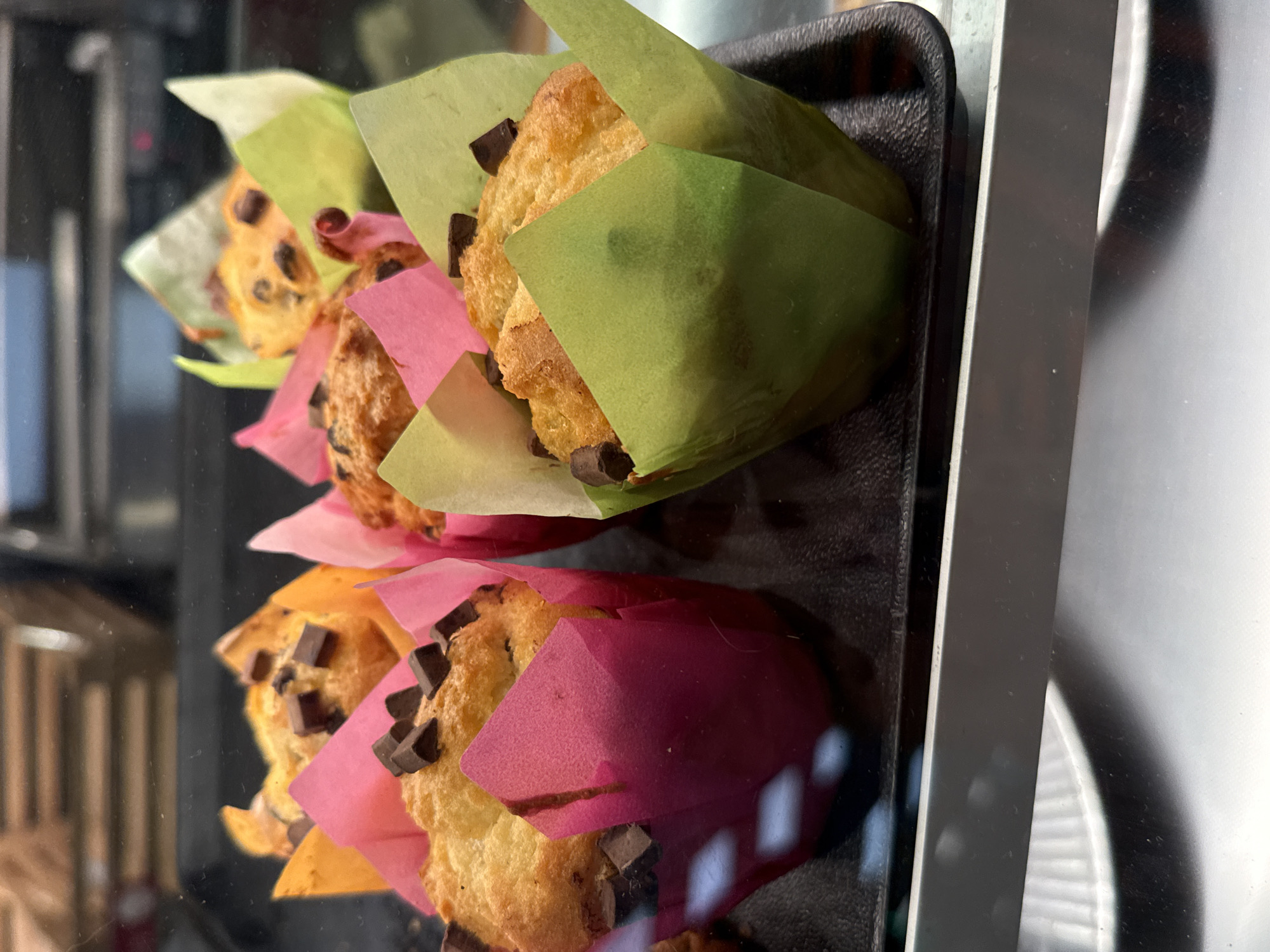 The image shows a close-up of several chocolate chip muffins in colorful paper liners, presented in a bakery display case. The muffins are golden brown with visible chocolate chips on top. They are arranged on a black tray, and the display case has a reflective surface. The background is blurred, showing what appears to be other baked goods or shelves in the bakery. The lighting suggests an indoor setting. This scene is consistent with a bakery in Herzberg, Germany, where such displays are common for fresh pastries.