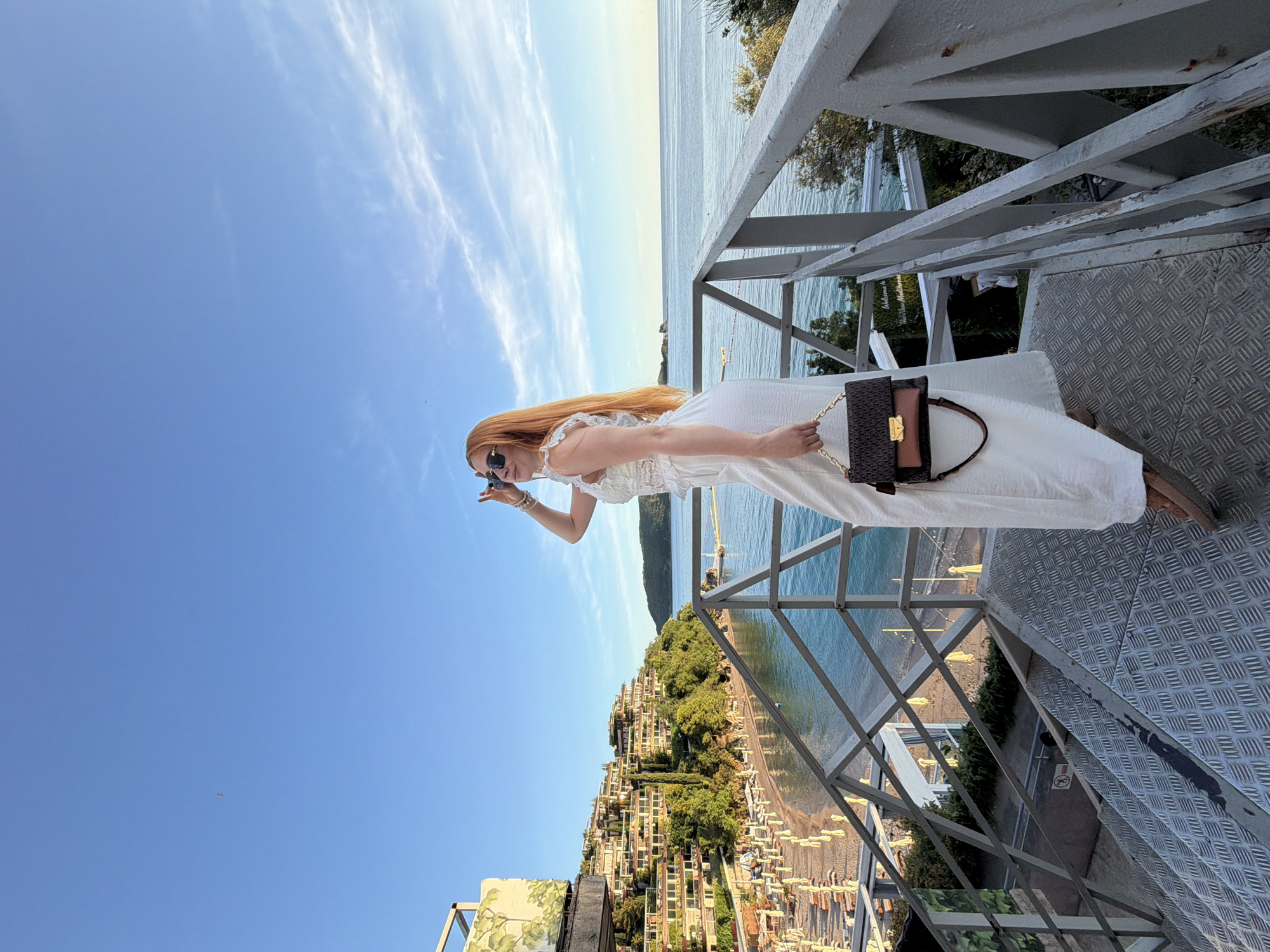 A bright, panoramic shot captures a woman standing on a metal viewing platform overlooking a stunning coastal landscape in Budva, Montenegro. The image, originally oriented vertically, is described here as if rotated correctly.
The subject is a woman with long, straight reddish-blonde hair, wearing a white, sleeveless dress or jumpsuit with delicate lace details. She wears dark sunglasses and holds a stylish dark brown and tan shoulder bag with a gold chain strap in her left hand. Her right hand is raised near her face, possibly adjusting her sunglasses or gesturing, as she gazes out at the extensive view.
She stands on a sturdy metal platform featuring a diamond-patterned floor and a white painted metal railing, which shows some signs of wear and rust. The railing extends across the upper right portion of the frame, providing a clear vantage point.
The background unfolds into a vibrant scene under a clear blue sky, dotted with a few wispy clouds. Below the platform, a dense hillside descends towards the sea, covered with numerous light-colored buildings, likely hotels or apartments, interspersed with lush green trees and vegetation. At the base of the hillside, a sandy or pebble beach is visible, lined with rows of sun loungers and umbrellas. The deep turquoise-blue water of the sea stretches out to the horizon, meeting the clear sky. Another distant landmass or mountain range can be seen across the water.
The scene depicts a beautiful sunny day, likely in the late morning or early afternoon, with calm weather. The woman appears to be enjoying the extensive scenic vista from this elevated viewpoint. Faint, indistinct text on a sign in the mid-ground, partially obscured by greenery, appears to read "Welcome Villa Meljine" or "Mali Meljine," providing a specific location cue within Budva.