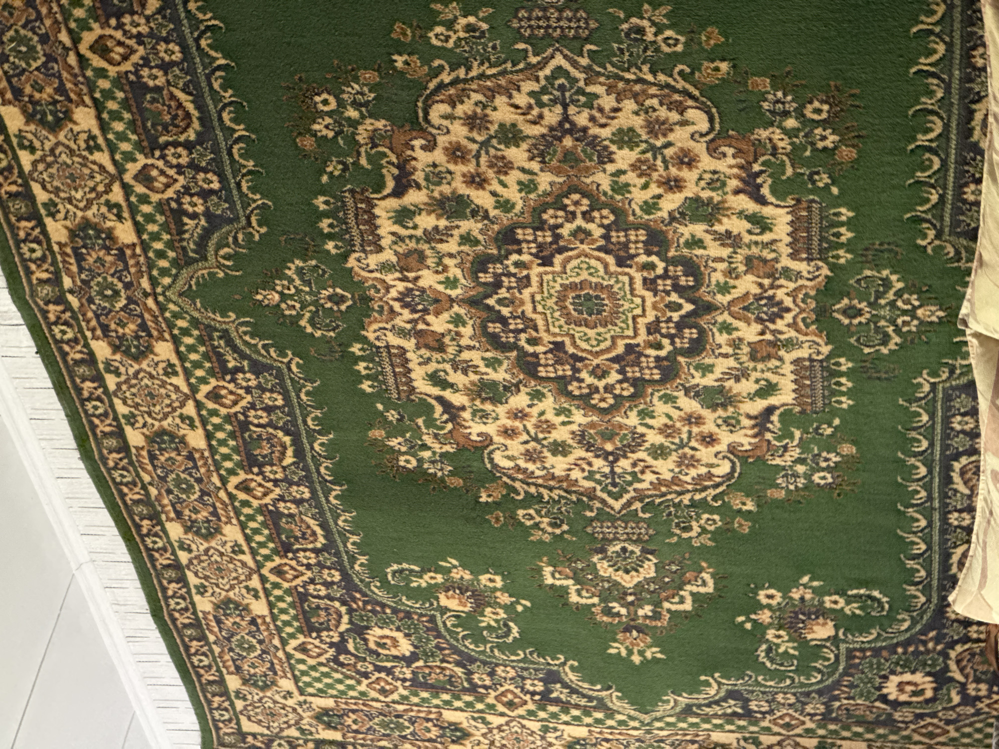 The image shows a close-up view of a large, intricately patterned Persian rug with a dominant green background. The rug features a central medallion design and floral motifs in colors such as beige, brown, and dark blue. The rug is hung vertically against a white, slatted wall, suggesting it might be displayed as a tapestry or covering. The top edge of the rug has a fringe. The lighting appears to be natural and diffuse, casting no harsh shadows. The bottom of the image shows the corner of what appears to be furniture with a patterned textile. There are no people or animals visible in the image. The location is identified as the city of Shymkent, Kazakhstan.