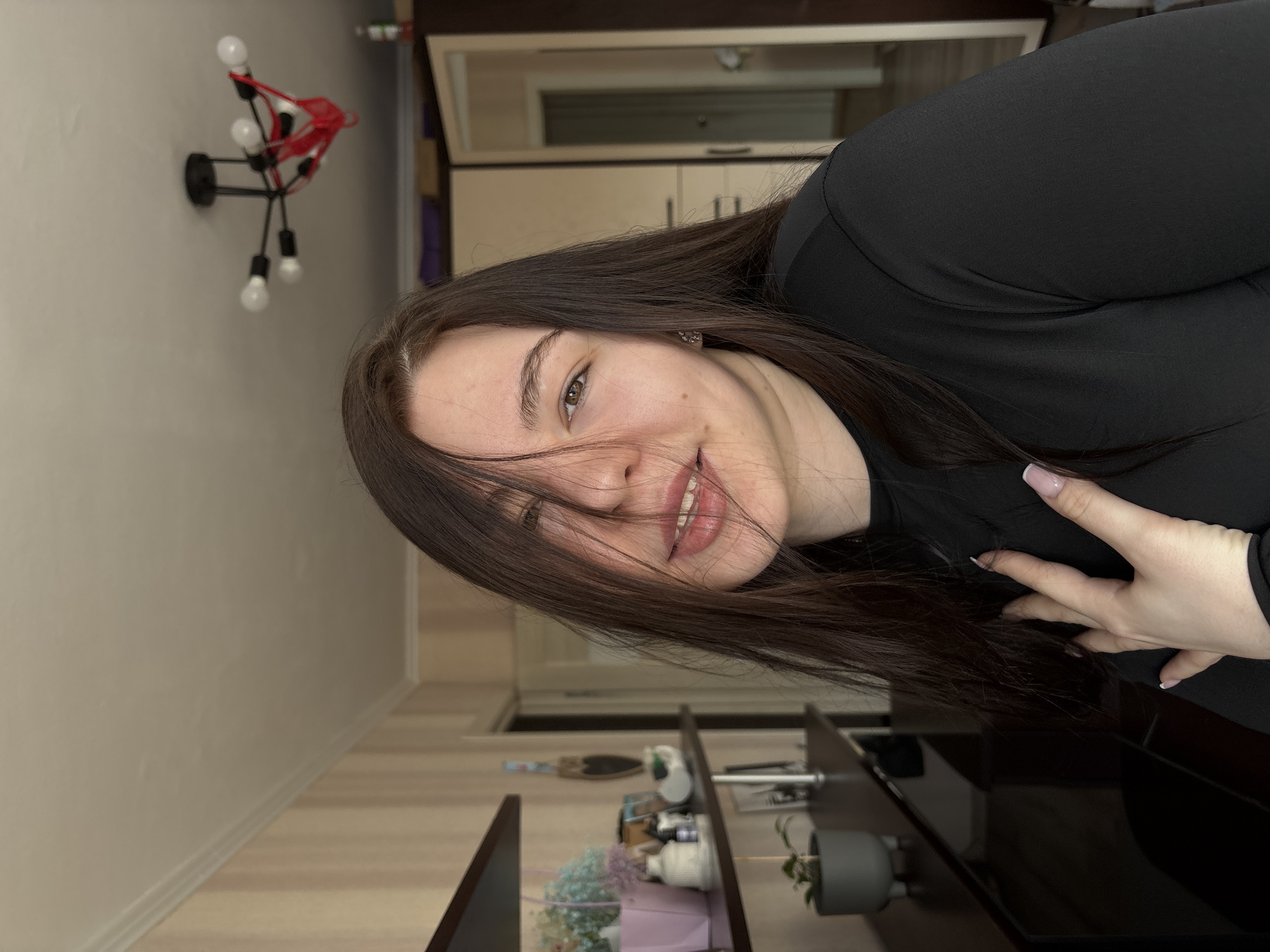 The image depicts a female subject in an interior residential setting in Perm, Russia. The subject is positioned centrally, viewed from the chest up, with long, dark brown hair partially obscuring the left side of her face. She has brown eyes and a subtle smile, with her teeth slightly visible. She wears a black, long-sleeved, crew-neck top. Her right hand, featuring light-colored, squared fingernails, is positioned with fingers spread on her upper chest or shoulder. A small silver stud earring is visible on her right earlobe.
The background on the left features vertical striped wallpaper in beige and light brown tones. A dark brown wall-mounted shelf holds a pink rectangular container with light blue and purple dried flowers, a white ceramic decorative object, a small dark bottle, and a framed photograph. Below this, a small green plant in a grey, footed pot rests on a dark, reflective surface, likely a cabinet or dresser.
The background on the right reveals part of a beige and dark brown wardrobe or cabinet, with one door slightly ajar, showing a glimpse of an interior space or another room. Above, a modern black ceiling light fixture with five exposed incandescent-style bulbs is visible, with a red thong-style undergarment suspended from one of its arms. The ceiling is white with crown molding.