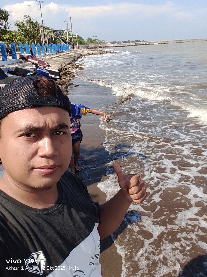 The image depicts a coastal scene in Limbangan, Indonesia, captured on October 12, 2025, at 14:16. A male individual, appearing to be an adult, is positioned in the immediate foreground, facing the camera. He wears a black mesh baseball cap backward and a black t-shirt with a white diagonal section and a grey graphic. His right hand is raised to chest height, forming a "thumbs up" gesture. Behind him, a second individual, partially visible from the torso down, stands in the shallow, brownish-green seawater on a dark sandy beach. This individual wears a blue and yellow short-sleeved shirt featuring an anime-style graphic and appears to be interacting with the water. Gentle, foamy waves break on the shore. Along the left side of the frame, a concrete seawall with blue painted railings extends into the distance, with erosion-control rocks visible at its base. Several small, overturned boat-like structures or debris are present on the sand next to the seawall. A utility pole is visible on the landward side of the seawall, with trees in the background. The sky is partly cloudy with blue patches, indicating bright daylight conditions.