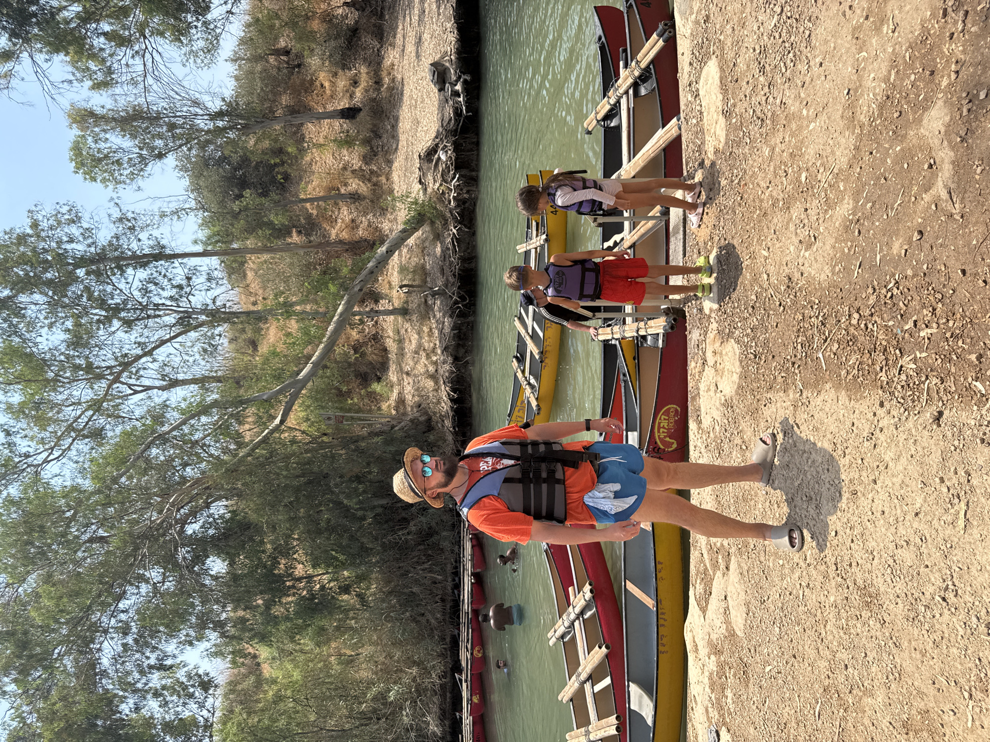 In Emek HaYarden Regional Council, Israel, a man wearing a straw hat, sunglasses, an orange t-shirt, a life vest, and blue shorts stands on a sandy bank next to a body of water. Canoes are lined up on the bank and in the water. In the background, several people are in the water, and trees line the opposite bank of the river. Two children, one boy and one girl, are standing on the bank near the man, also wearing life vests. The boy is wearing red shorts and the girl is wearing a white swimsuit. Another adult, partially visible, stands behind the children. The river is a light green color, and the day appears to be sunny and warm.