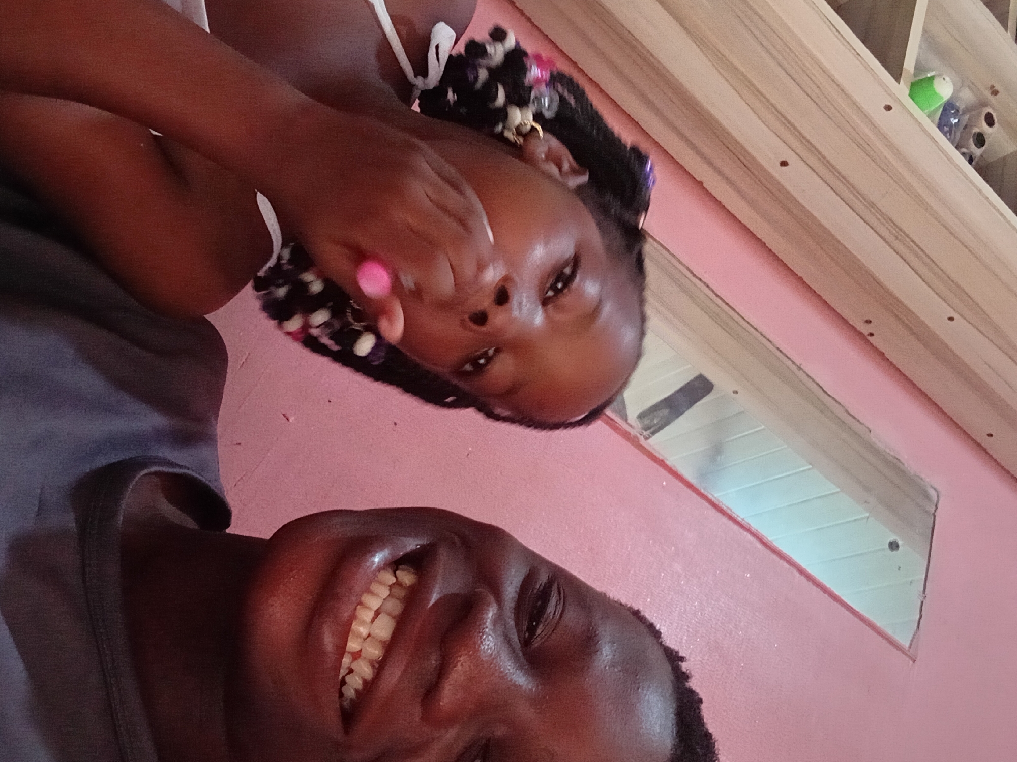 Two children, a girl and a boy, are captured in a close-up selfie. The girl, on the left, has dark skin and braids adorned with colorful beads. She is wearing a white top and holds a pink object, possibly candy, near her mouth. Her gaze is directed towards the camera. The boy, on the right, also has dark skin and is smiling broadly, revealing his teeth. He is wearing a gray t-shirt. The background consists of a pink wall and a wooden shelf unit with some items on it. A partial reflection of the room is visible in a mirror on the wall. The image appears to be taken indoors in what seems to be a domestic setting.