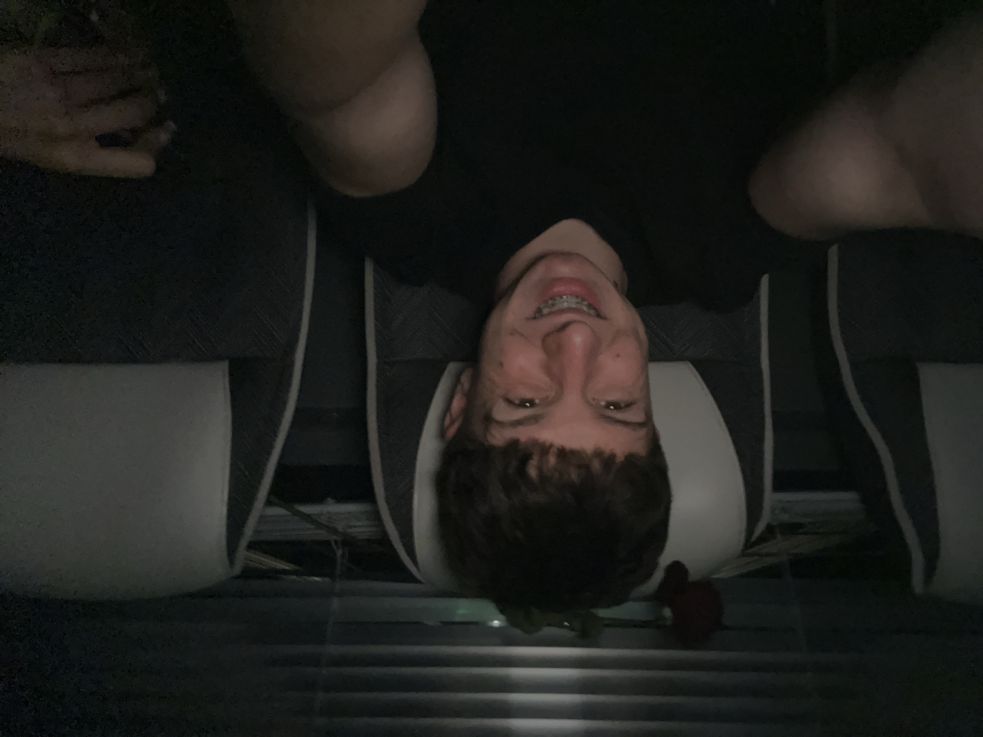 A male individual with brown hair is lying down, seemingly supine, in what appears to be a car. The man is looking up with a smile, displaying dental braces. A red rose is situated near his head, possibly on the floor of the vehicle. The interior of the vehicle features gray and white seats. The location cannot be determined from the image.