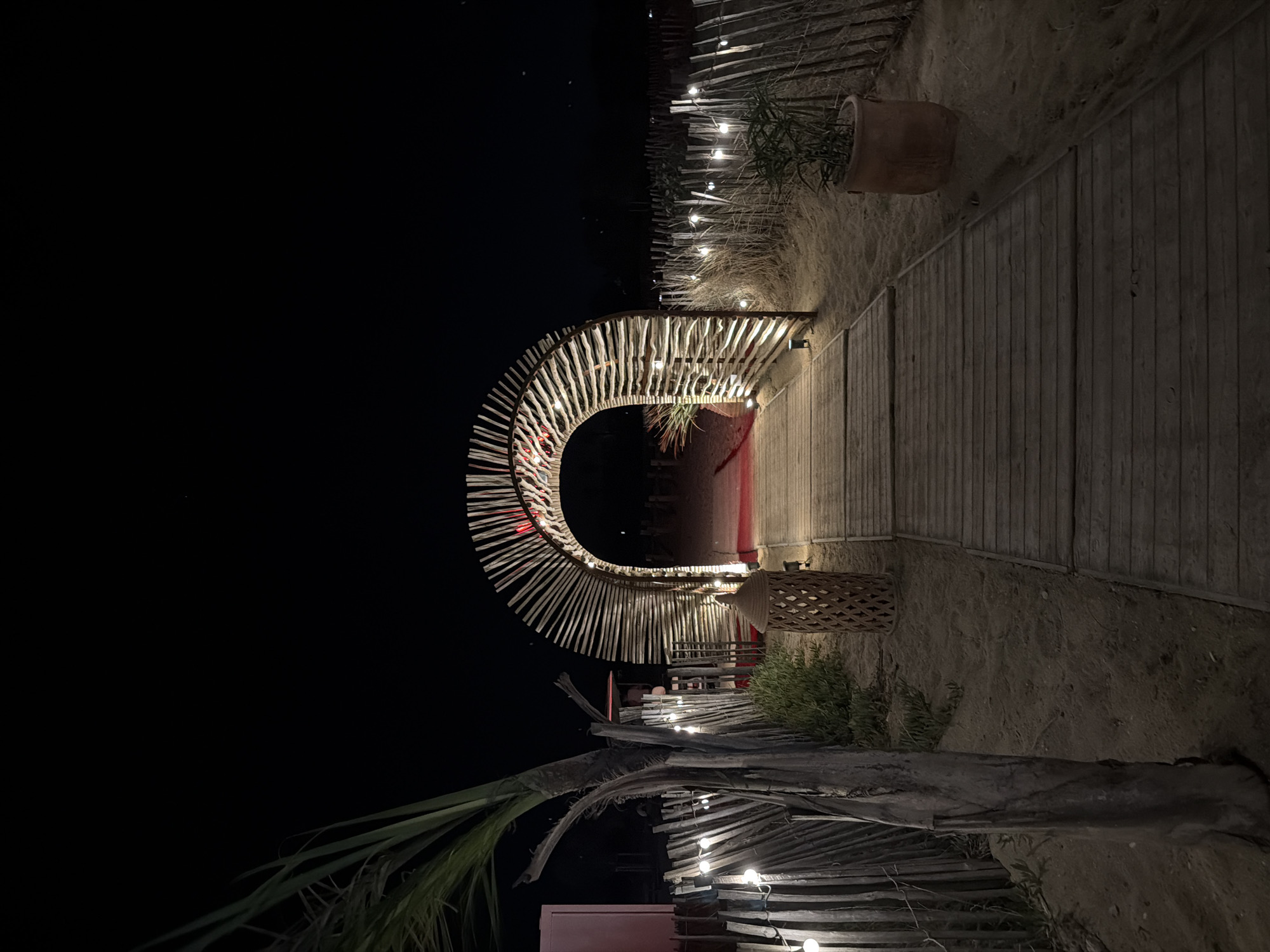 The image depicts an illuminated archway made of natural wood slats on a sandy beach at night, located in Torreilles, France. A wooden boardwalk leads through the arch, which is adorned with string lights. To the left, a palm tree stands beside a rustic wooden fence also decorated with string lights. A woven lantern sits on the sand near the boardwalk, and a potted plant is visible to the right. The background is dark, suggesting nighttime, with a dimly lit structure visible through the arch. The overall scene conveys a festive and inviting atmosphere.