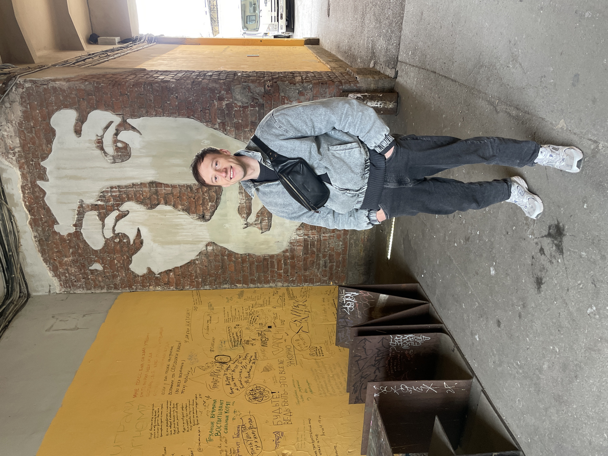 A man stands in an urban passage or alleyway, looking directly at the camera with a cheerful smile. He has short brown hair and is wearing a light grey denim jacket, a dark grey top with ribbed cuffs, black jeans, a black fanny pack worn across his chest, and light grey and white athletic shoes. He stands with his hands in his pockets, appearing relaxed.
The passage features a distinctive brick wall on the left, adorned with a large, partially eroded mural of a stylized face, rendered in white and exposed brick, possibly depicting John Lennon. Below this mural, the wall transitions to a bright yellow surface densely covered in handwritten messages, drawings, and graffiti in various languages, predominantly Russian. Some visible phrases include "играли," "Мне голос был он звал утешно," "Я люблю НАТАШУ!", "#Сюсинг," "Привет Аркади," "Трудные времена воспитывают сильных котят" (Hard times raise strong kittens), "БУДЬТЕ!", "ВЕДЬ БЫТЬ - ЭТО ВСЕ ХЕ ЗАКОННО" (Because to be is all lawful), and "VANCH."
The ground is a worn concrete pathway. On the right side, there are abstract, multi-tiered metal structures or steps, also covered in graffiti. The passage opens up overhead to a bright, clear sky, indicating it is daytime. In the distance, a white truck is visible, with part of its license plate "X789CT | 99" discernible. The scene presents a vibrant, gritty, and artistic urban environment, characteristic of places like Saint Petersburg, Russia, where this photo was taken.