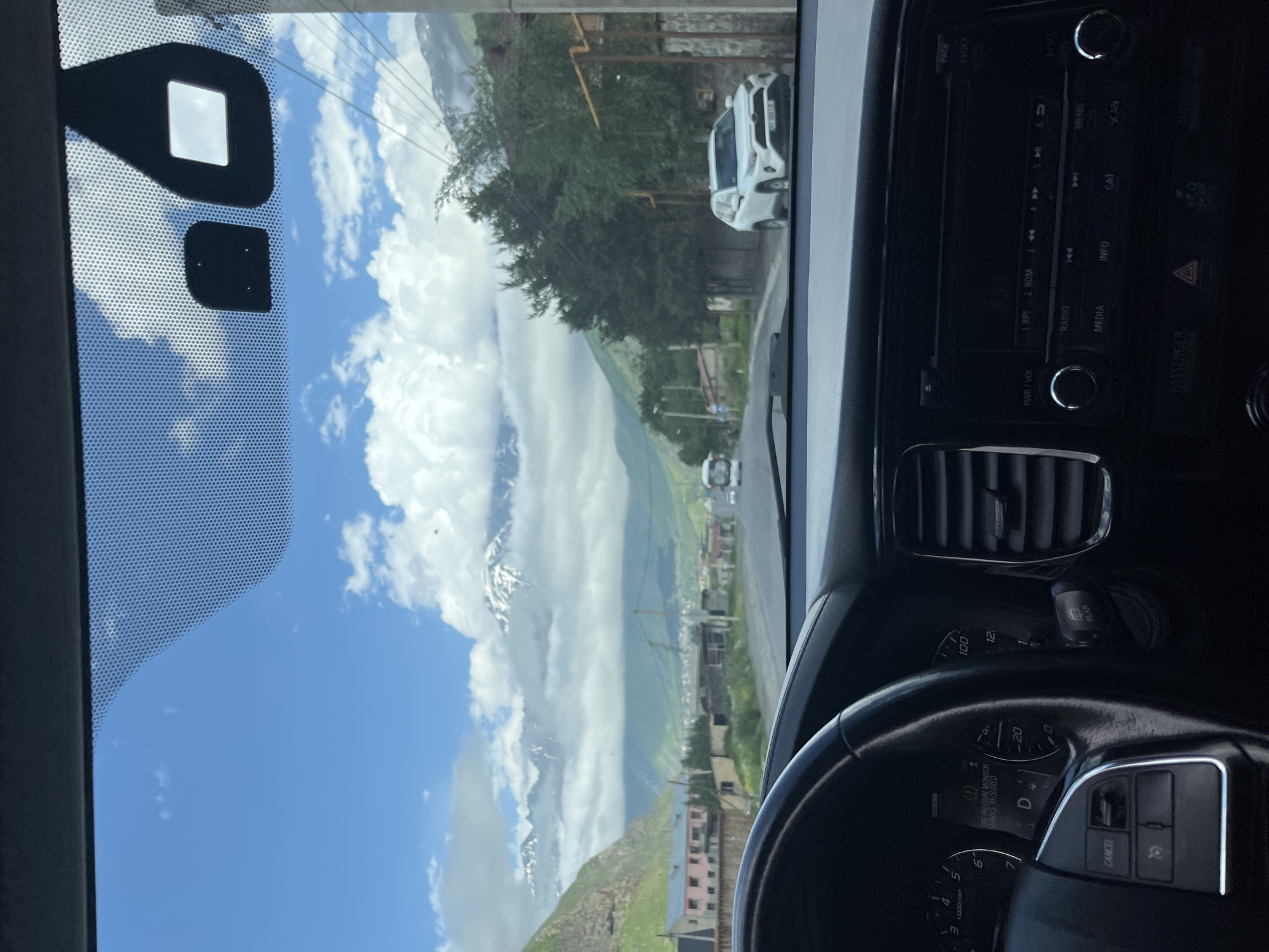 The image is taken from inside a car, looking out through the windshield. The view shows a road in a mountainous area. The road curves to the right, with trees and buildings lining its sides. In the distance, snow-capped mountains rise against a blue sky with scattered white clouds. A white SUV is parked on the right side of the road. The interior of the car is visible, including the steering wheel, dashboard with a digital display showing "TIRE PRESSURE MONITOR SERVICE REQUIRED", and the car's entertainment system. The location is identified as Arsha, Georgia. The scene depicts a journey through a scenic landscape.