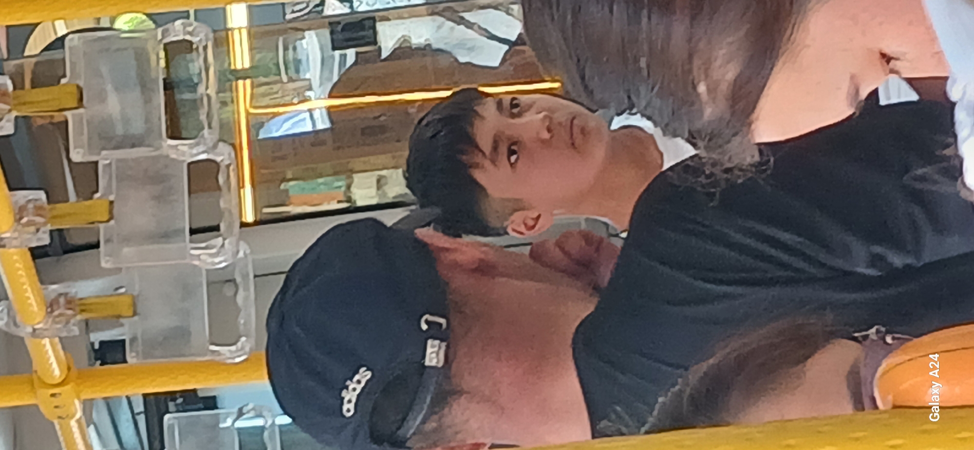 The image shows the interior of a bus with yellow handrails and clear plastic grab handles. In the foreground, a person wearing a dark baseball cap with an Adidas logo is partially visible from behind, facing away from the camera. To their right, a young person with short, dark hair is looking towards the camera with a neutral expression. Their attire includes a white t-shirt. In the background, a reflection in what appears to be a window or mirror shows a blurred scene of buildings or structures. The lower right corner of the image contains the text "Galaxy A24".