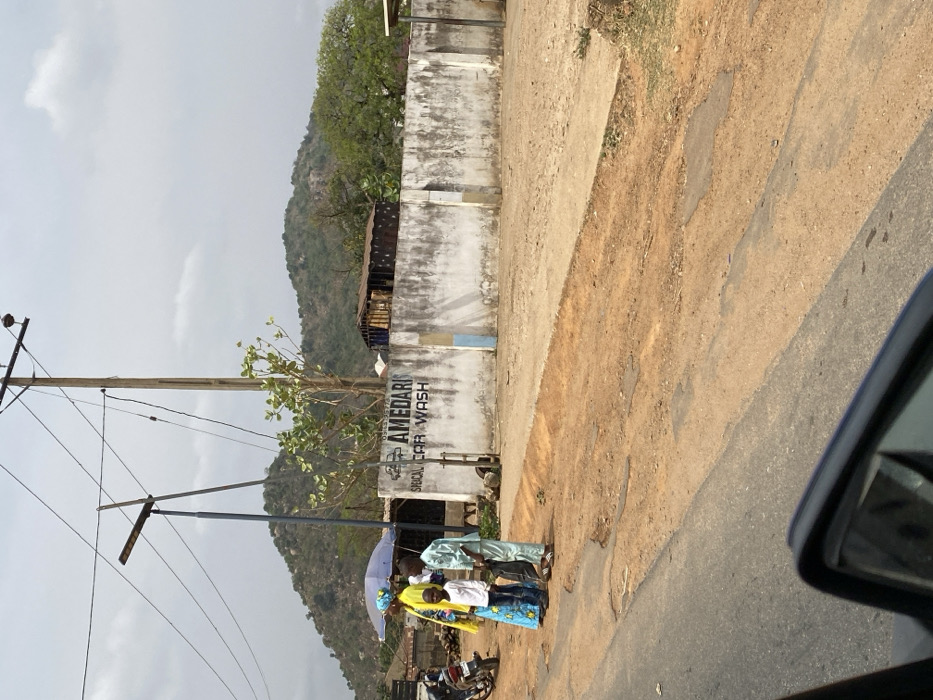 The scene depicts a roadside in Magongo, Nigeria, viewed from the interior of a vehicle with a portion of its side mirror visible in the lower right. An asphalt road with visible wear transitions to a dirt shoulder. On this shoulder, three individuals are present: an adult female in a yellow top and blue patterned skirt, holding a light blue umbrella; a child wearing a white shirt and blue jeans; and a second child in a light blue tunic and trousers. Behind these individuals, a stained white concrete wall displays the black lettering "AMEDARIS SPECIAL CAR WASH," with a partial phone number "0913-..." above. Two poles rise vertically behind the wall: a wooden utility pole carrying multiple wires and a narrower, darker pole supporting a solar street light. A small, dilapidated structure with a rusty corrugated metal roof and exposed framework is visible behind the wall to the right. A motorcycle is parked on the far left of the scene. The background features several rocky, tree-covered hills under a hazy, overcast sky.