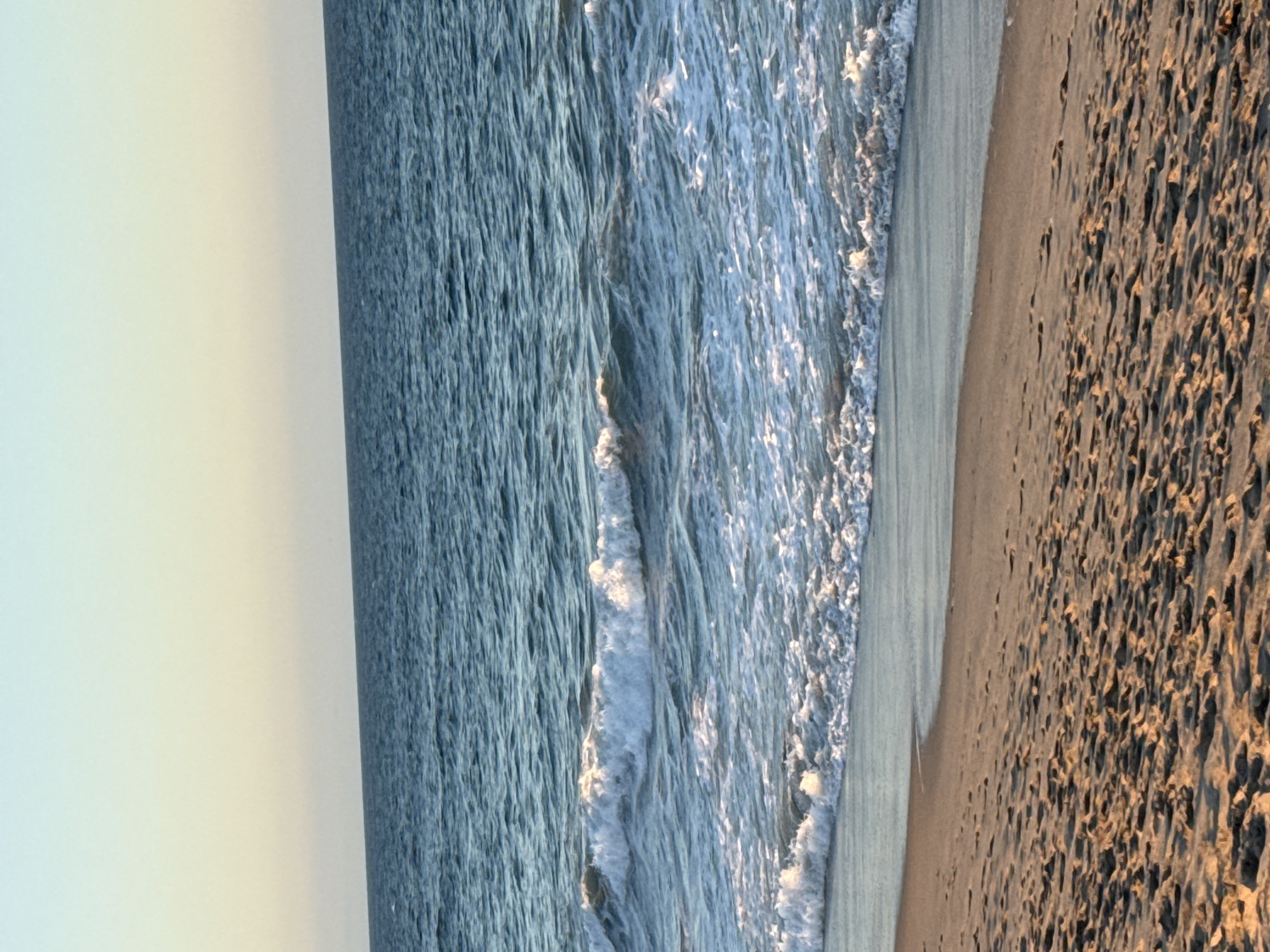 The image depicts a tranquil beach scene at dusk in Malibu, California. The foreground is dominated by soft, sun-kissed sand, imprinted with numerous footprints, suggesting recent human activity. The sand transitions to the gentle, lapping waves of the Pacific Ocean. The water exhibits a deep blue hue, with white foam cresting on the incoming waves, creating a dynamic yet soothing visual. The horizon is a sharp line where the ocean meets a pale, gradient sky, transitioning from a soft yellow near the horizon to a clear, light blue at the top of the frame. There are no people or specific landmarks visible, emphasizing the natural beauty of the coastline.