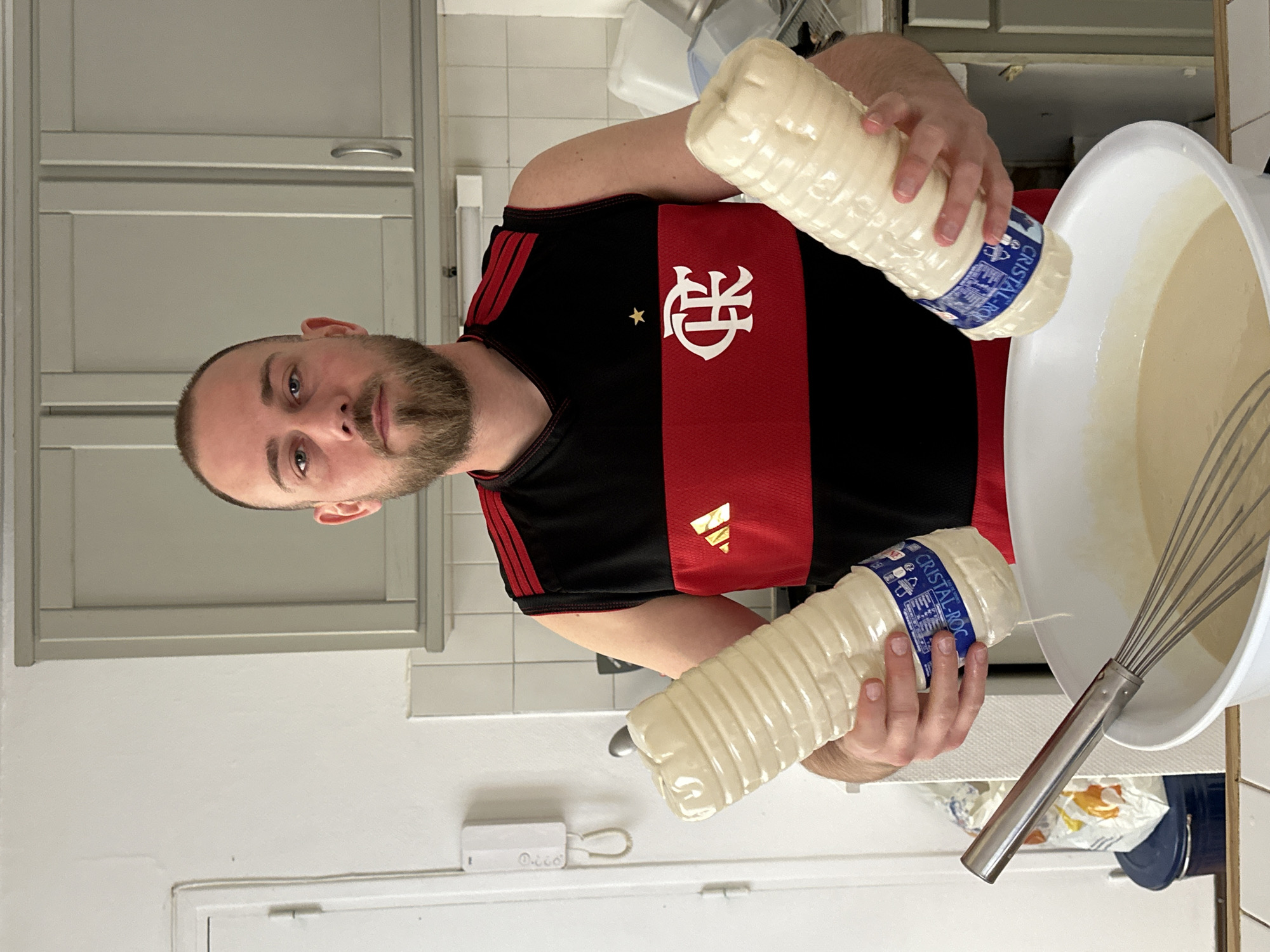 A man in a Flamengo jersey is preparing food in a kitchen. He is holding two bottles labeled "CRISTAL-ROC" and pouring the contents of one bottle into a white bowl. A whisk is also in the bowl. The kitchen has white cabinets and white tiled walls. The location is likely Paris, France, based on the brand of the bottle being a local dairy brand.