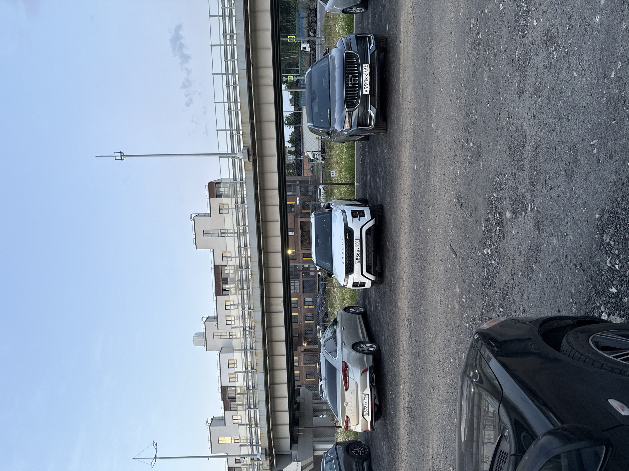 The image shows a parking lot filled with several cars. In the foreground, a black car's front right fender and hood are visible. Behind it, a silver Hyundai Sonata is parked, displaying a license plate that reads "B672TK 790". To its right is a white Range Rover Evoque, with a license plate "E856BY 250". Further to the right is a dark gray Geely Monjaro, its license plate visible as "E991KK 797". In the background, there is a large elevated structure, likely a bridge or highway overpass, with modern buildings behind it. The sky is partly cloudy with a pale blue hue. The ground is covered in gravel. The scene is set in a developed urban or suburban area, consistent with the city of Voyennosluzhashchiy, Russia. There are no people or animals visible in the image.
