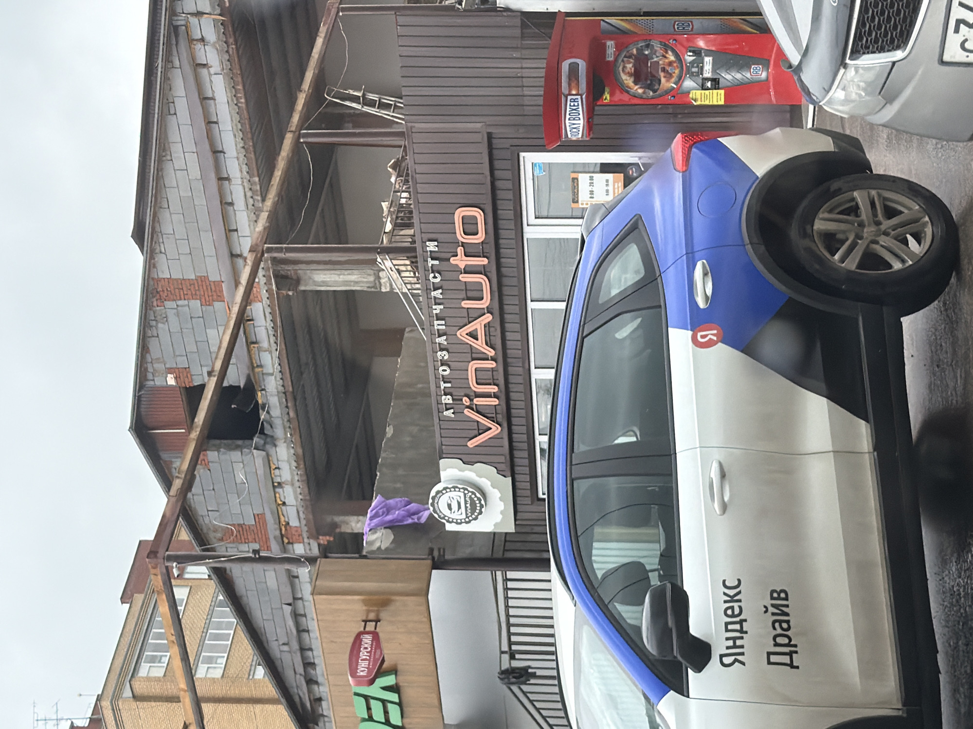 This is a vertically oriented, outdoor photograph taken in Фролы, Russia, likely during the day under overcast skies, as suggested by the diffused light and the wet pavement. The primary focus is on a white car with blue and grey accents, part of a ride-sharing service, parked in front of a commercial building. The car's side displays text in Russian, "Яндекс Драйв" (Yandex Drive), indicating its affiliation.
The building behind the car appears to be under renovation or in a state of disrepair. It has a dark brown, corrugated metal facade with a prominent sign that reads "VinAuto АВТОЗАПЧАСТИ" (VinAuto Auto Parts) in large, orange, illuminated letters. Below this, a circular logo with a vehicle silhouette is visible, also with "VinAuto" inscribed. A red vending machine, labeled "ROCKY BOXER" and with "RB" logos, is situated to the left of the "VinAuto" sign. Further to the left, a sign with green and red lettering, partially visible, reads "КУНГУРСКИЙ" (Kungurskiy), which likely refers to a local product or business.
The scene suggests a commercial area, possibly a street or parking lot, with elements of commerce and transportation. The wet appearance of the ground implies recent rain. No people are clearly visible, though shadows suggest some presence within the building or on the street. The overall impression is one of everyday urban activity, with a focus on vehicles and local businesses.