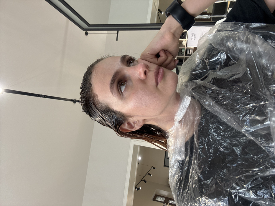 A female individual is depicted in a hair salon in Tbilisi, Georgia, undergoing a hair treatment. Her dark brown hair is wet and coated with a light brown substance, likely hair dye or a conditioning agent. She is wearing a clear plastic protective cape over a dark garment, covering her shoulders and chest. Her right hand is positioned under her chin, supporting her head, and she wears a dark, wide wristband on her left wrist. Her gaze is directed upwards and to her right, away from the camera, with a pensive expression. The immediate environment includes a white ceiling with a black track light fixture featuring a circular spotlight overhead. In the background to the right, a large, black-framed mirror reflects the room, while shelving stocked with various products and a partial "Drinks menu" sign are visible beneath it. To the left, an open doorway or archway leads to another area of the salon, where additional track lighting and a multi-paned arched window are partially visible.