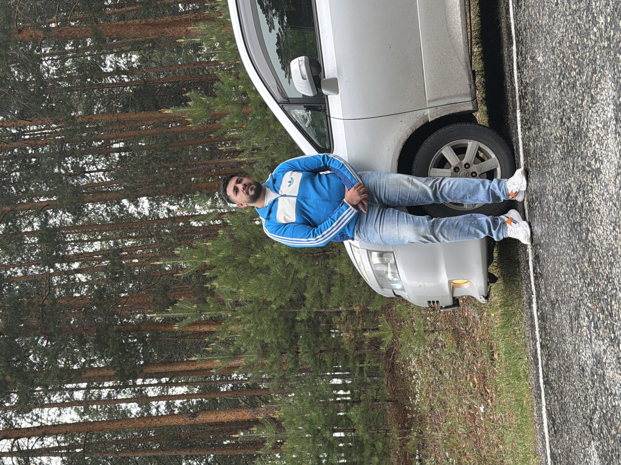 A man wearing a blue and white track jacket and jeans is leaning against the driver's side of a silver van parked on the shoulder of a road in a wooded area. The road is asphalt with a white line marking the edge. The trees in the background appear to be pine trees, indicating a forest-like environment. Based on the provided location of Сизая, Russia, the image likely represents a roadside scene in this region. The interaction is between the man and the vehicle in a natural environment near a road.